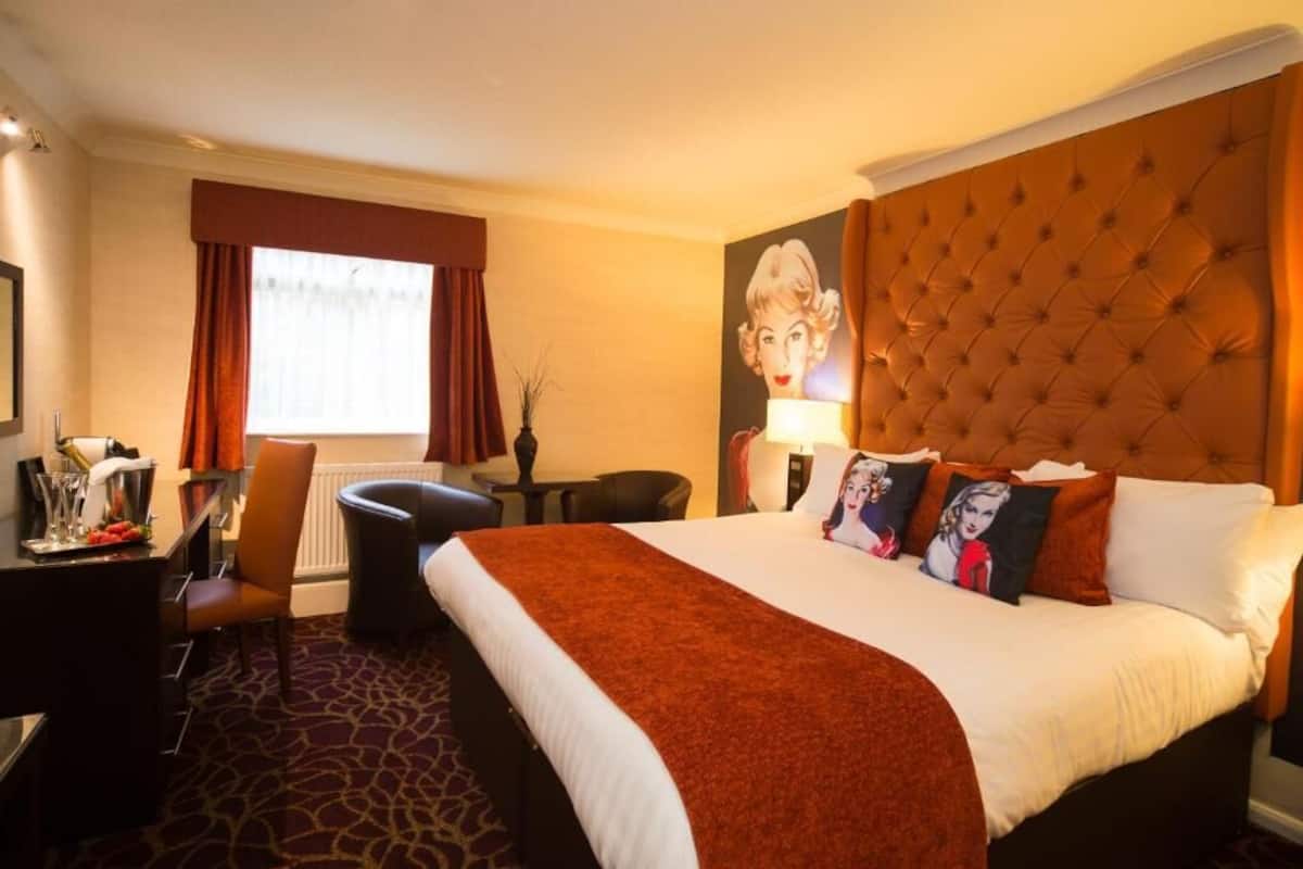 A hotel room with a large bed, a desk with a chair, a window with curtains, and a wall-mounted lamp.