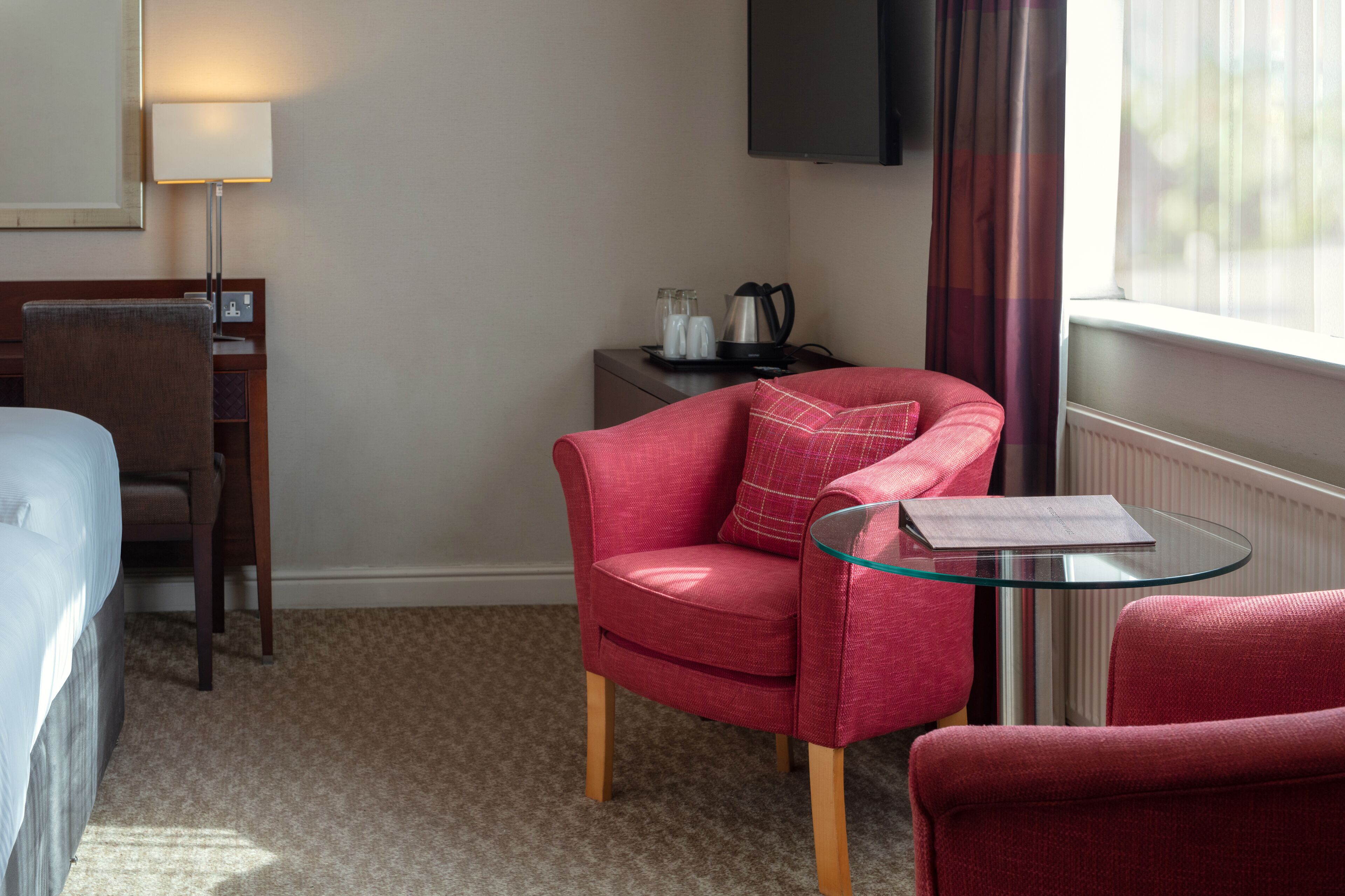 Habitación de hotel con cama, un sillón rojo, una mesa de vidrio, un escritorio con lámpara, televisor y ventana con cortinas.