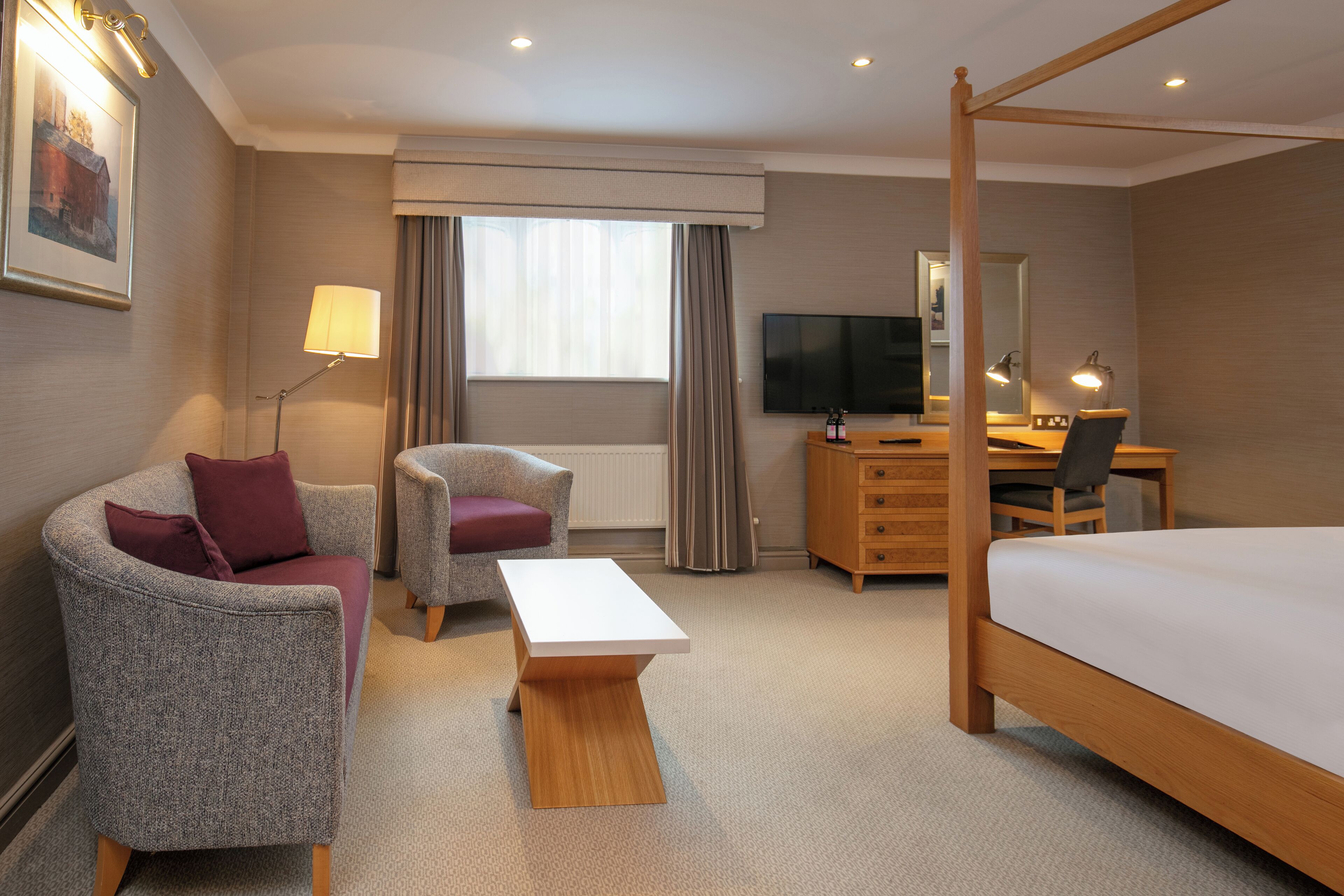 A hotel room with a four-poster bed, a desk with a computer, a television, and two armchairs.