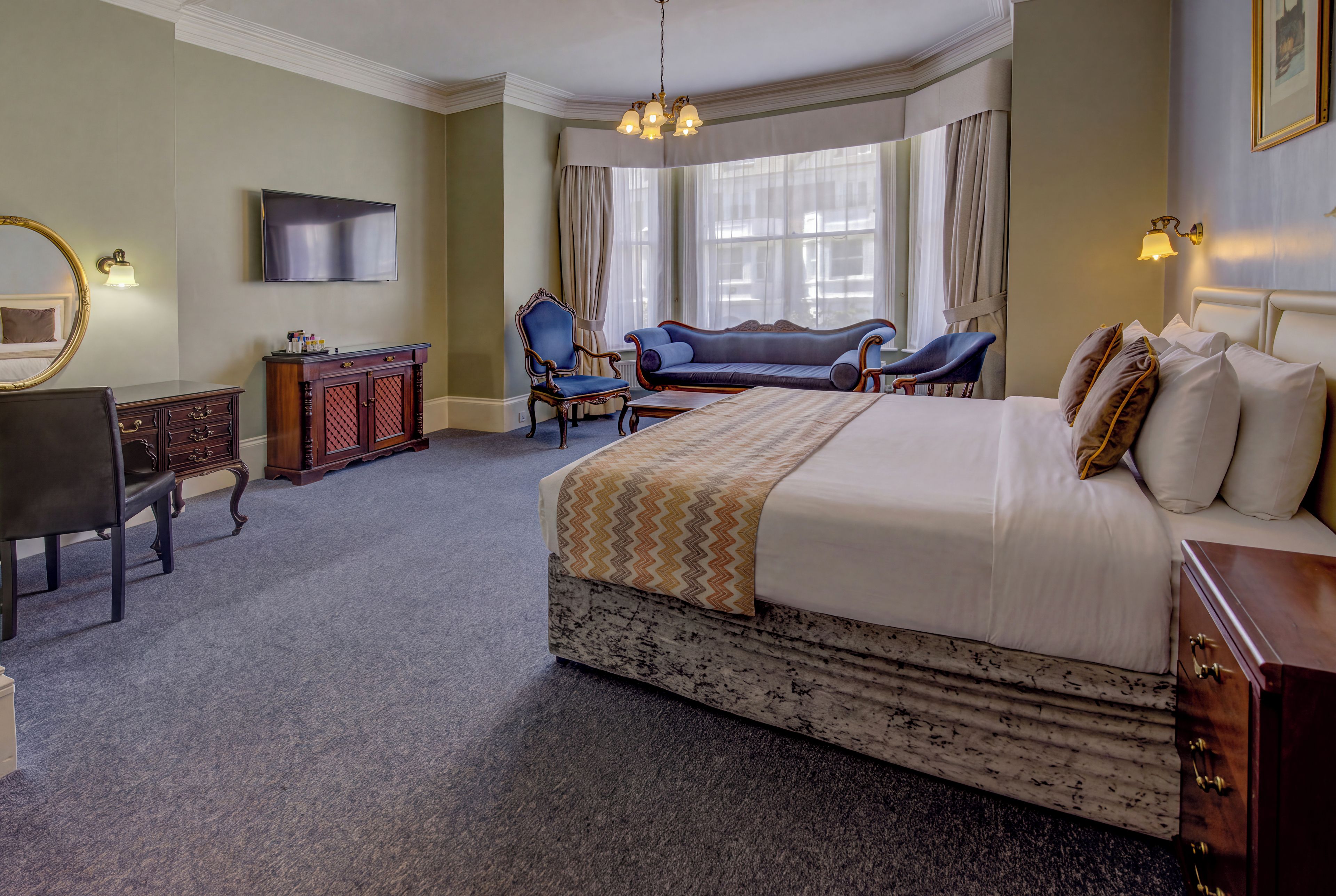 A hotel room with a large bed, a desk, a chair, a sofa, and a television.