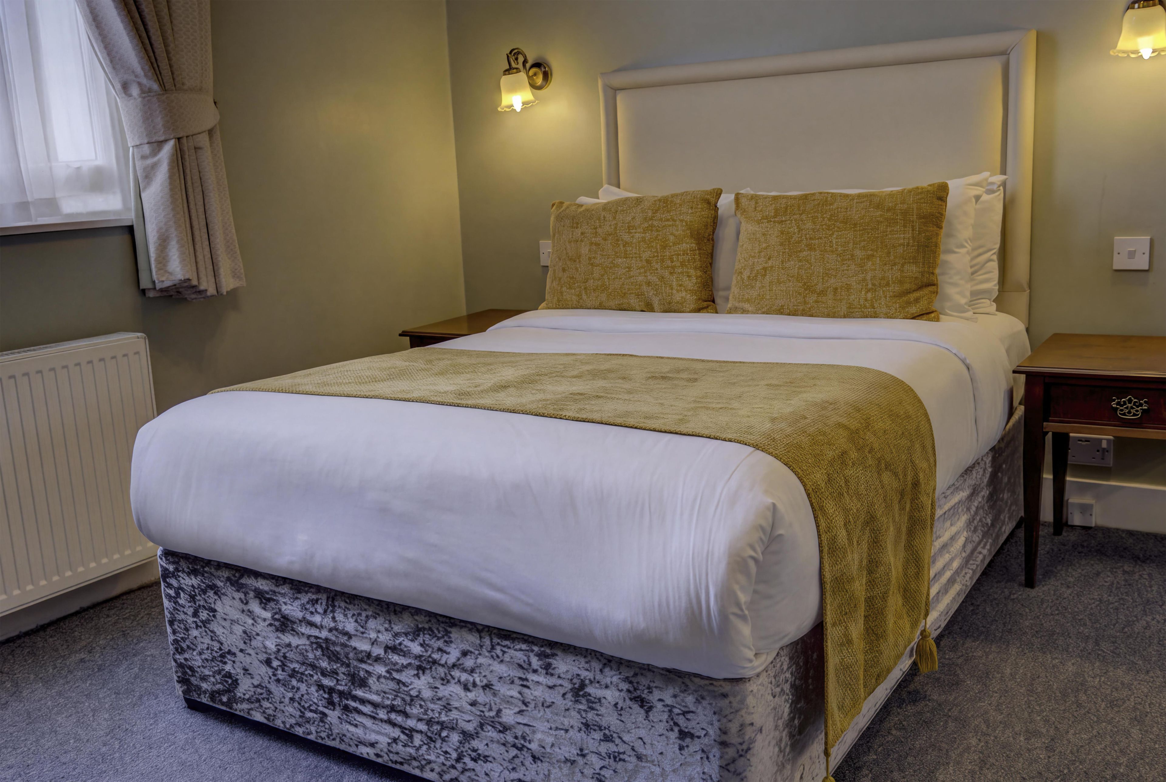 A neatly made bed with a white duvet and a beige throw blanket, a bedside table with a lamp, and a wall-mounted light fixture.