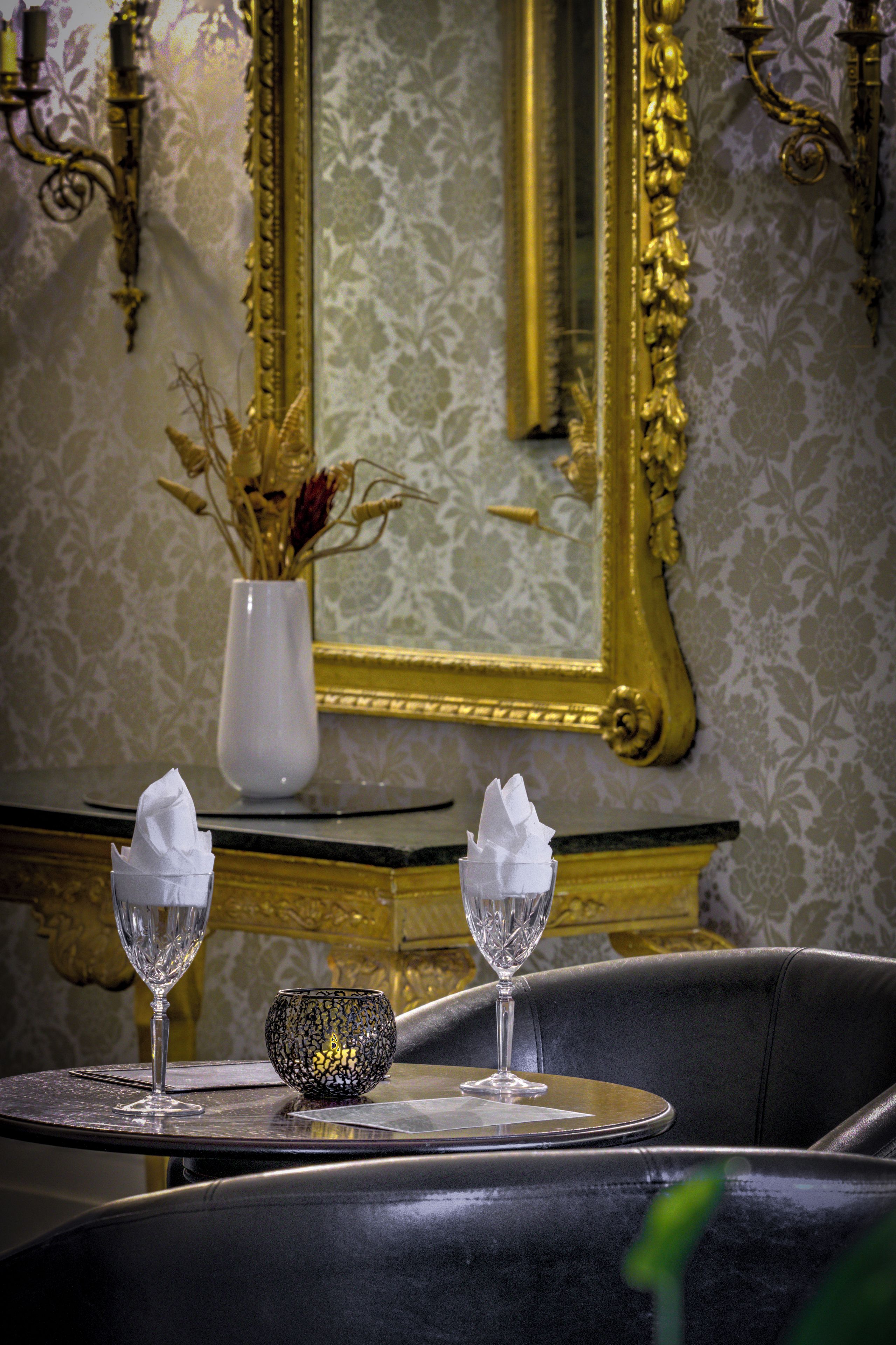 Una mesa de comedor con dos copas de cristal y un cuenco decorativo, frente a un espejo y ornamentos en la pared.