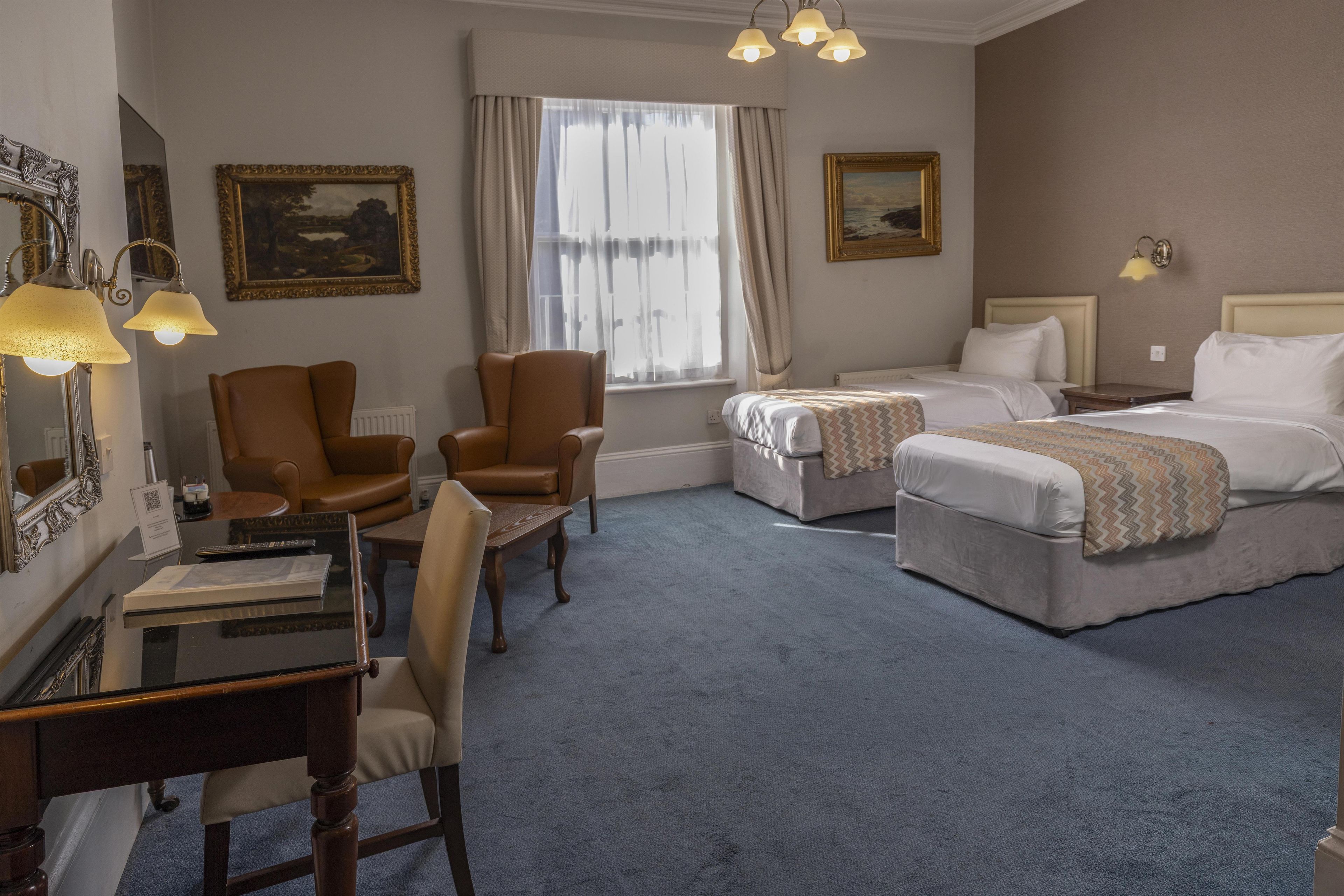 A hotel room with a desk, two chairs, a bed, and a window with curtains.