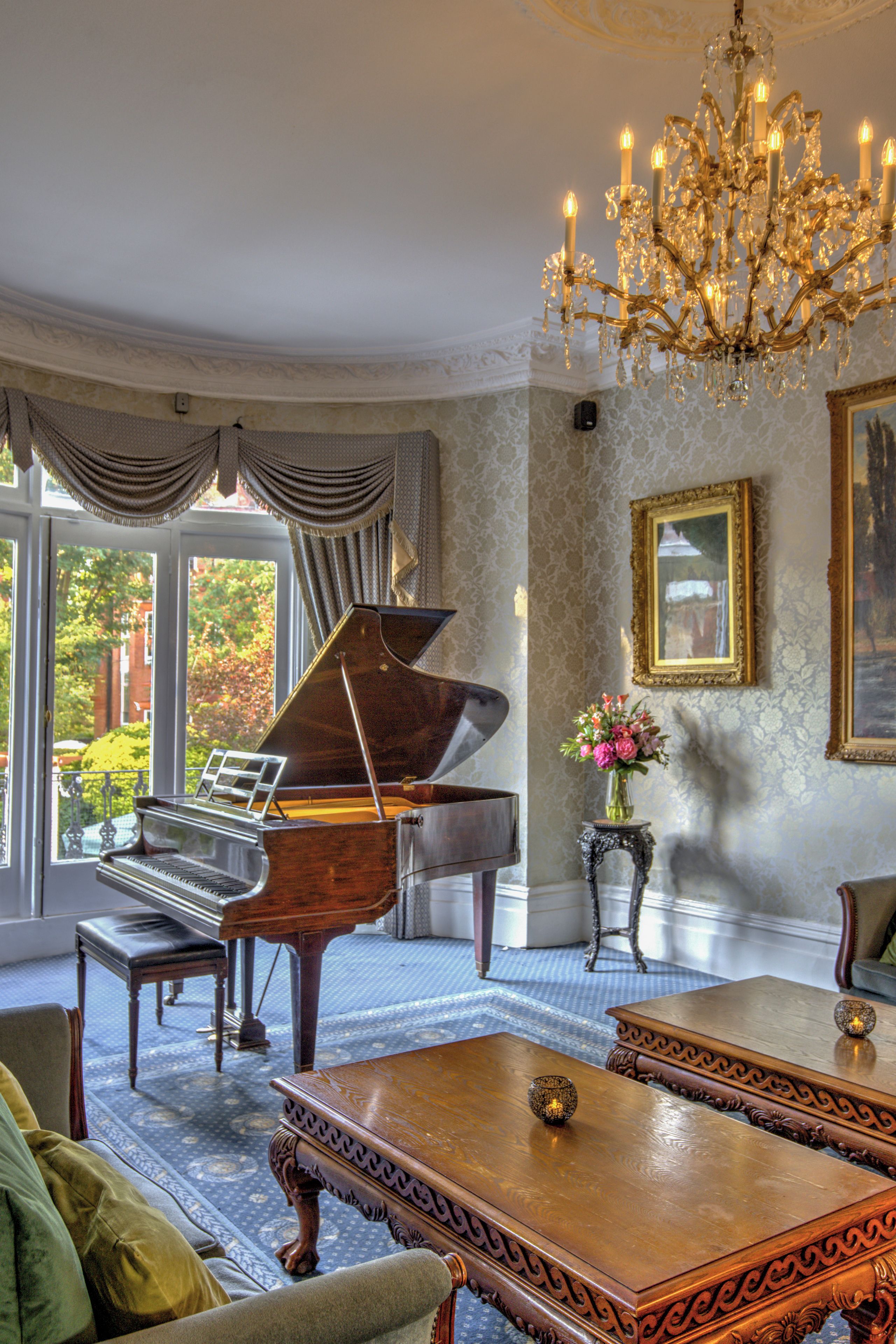 A grand piano in a room with a chandelier, a painting, and a view through a window.