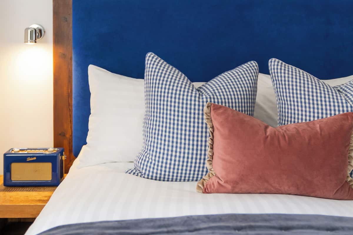 A bed with a blue upholstered headboard, checkered pillows, a pink pillow, and a radio on the nightstand.