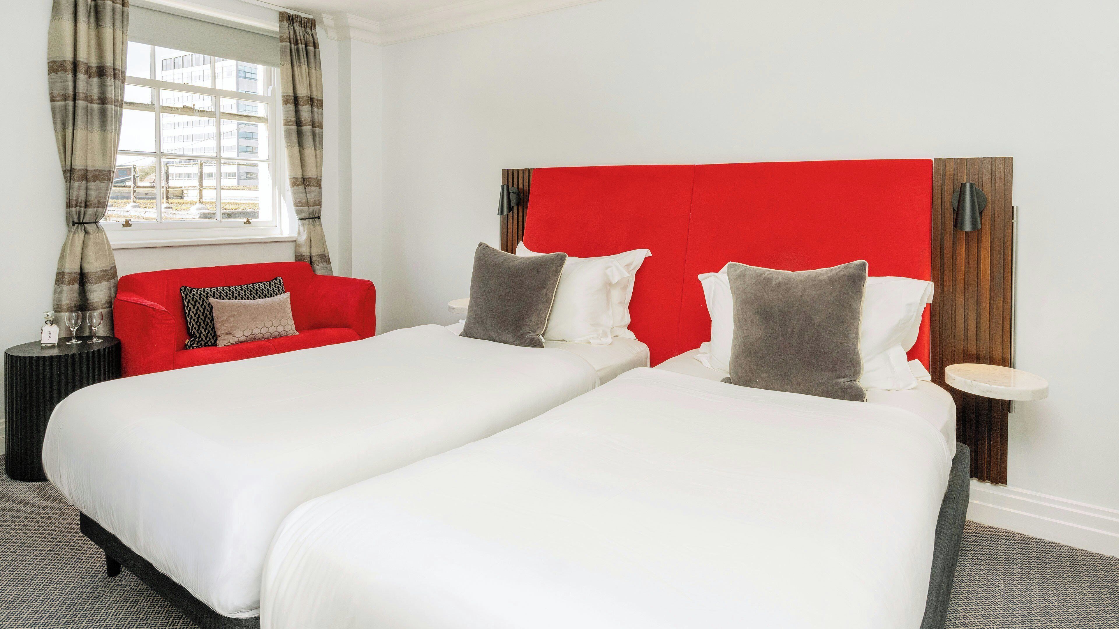 A hotel room with two beds, a red headboard, a red sofa, and a window with curtains.