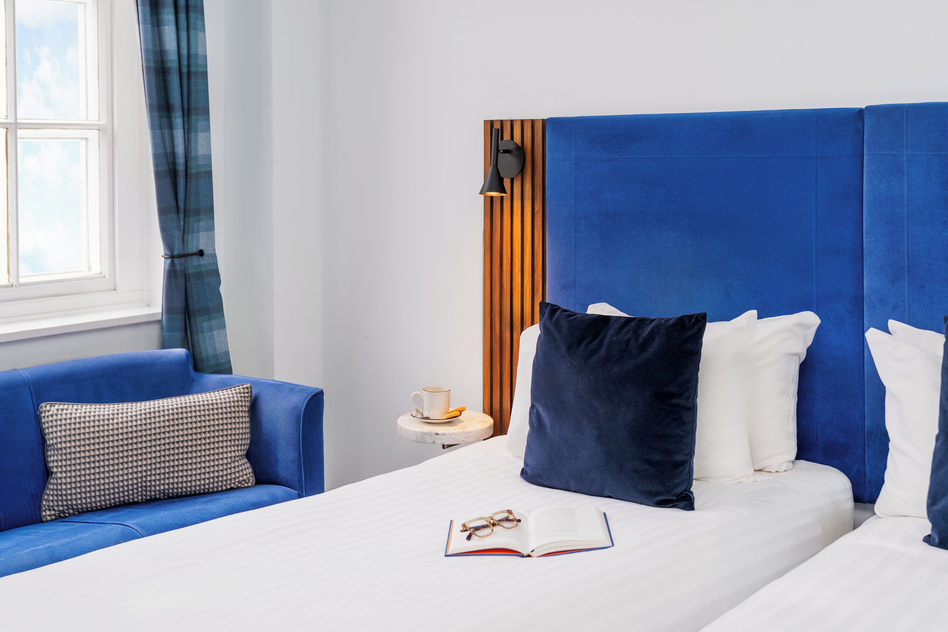 A hotel room with a large bed, a blue headboard, a bedside table with a cup and saucer, and a blue sofa with patterned cushions.