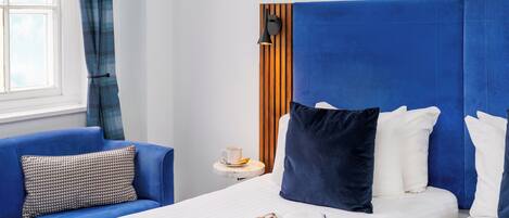 A hotel room with a large bed, a blue headboard, a bedside table with a cup and saucer, and a blue sofa with patterned cushions.