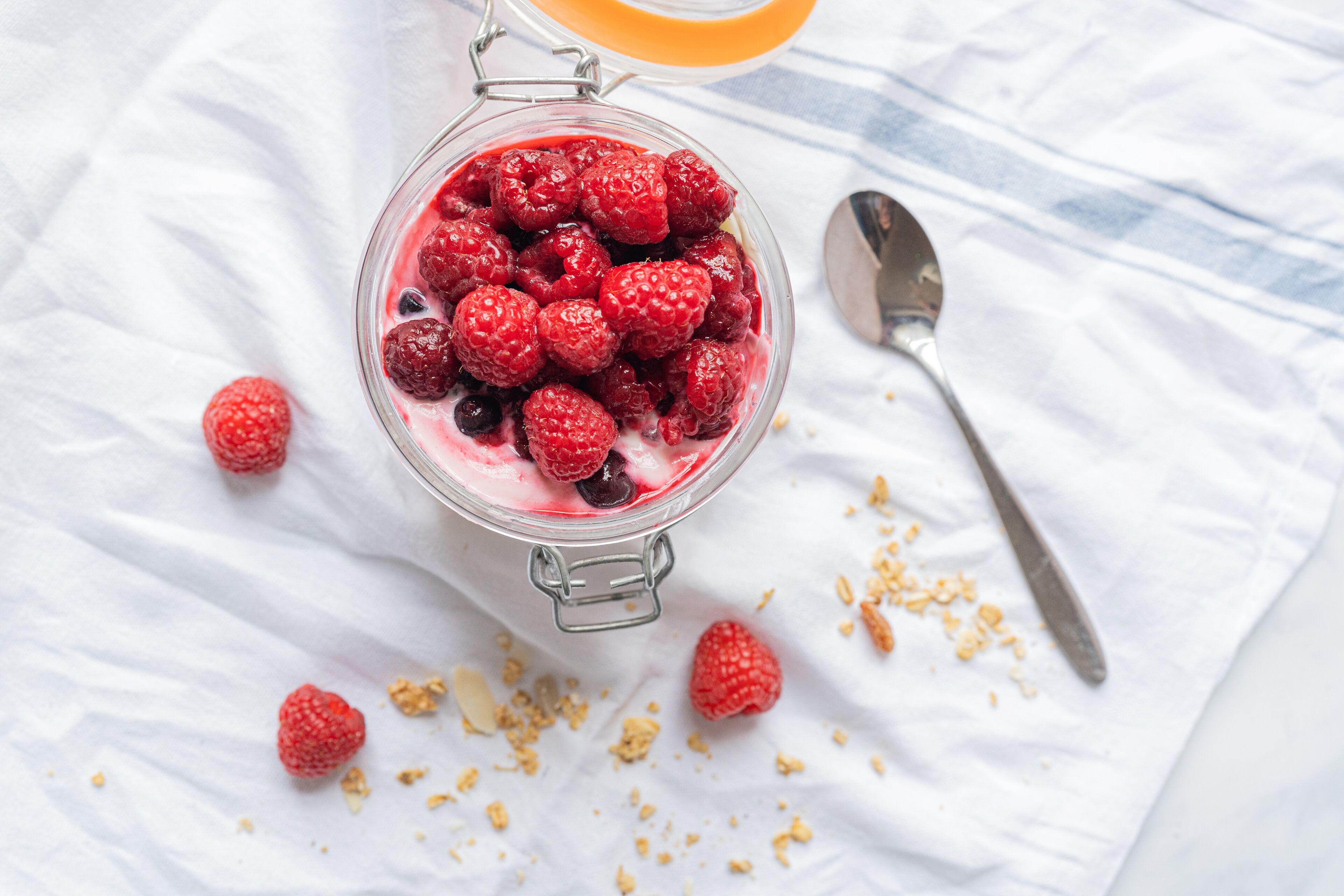 Ein Glasgefäß, gefüllt mit Himbeeren, und ein Löffel liegen daneben auf einer weißen Tischdecke.