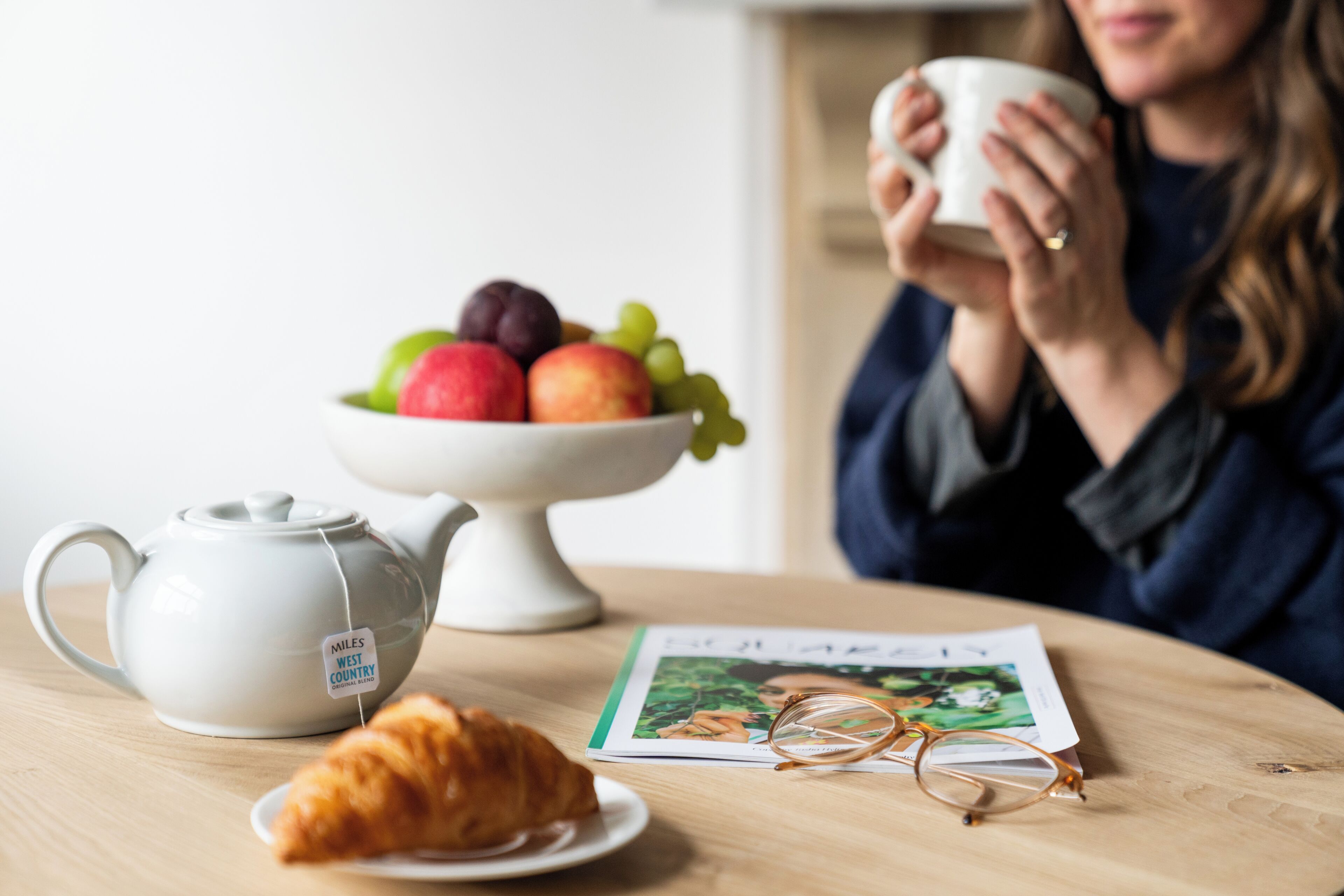 Una mesa con una tetera, un bol de fruta, un croissant, un par de gafas y una revista.