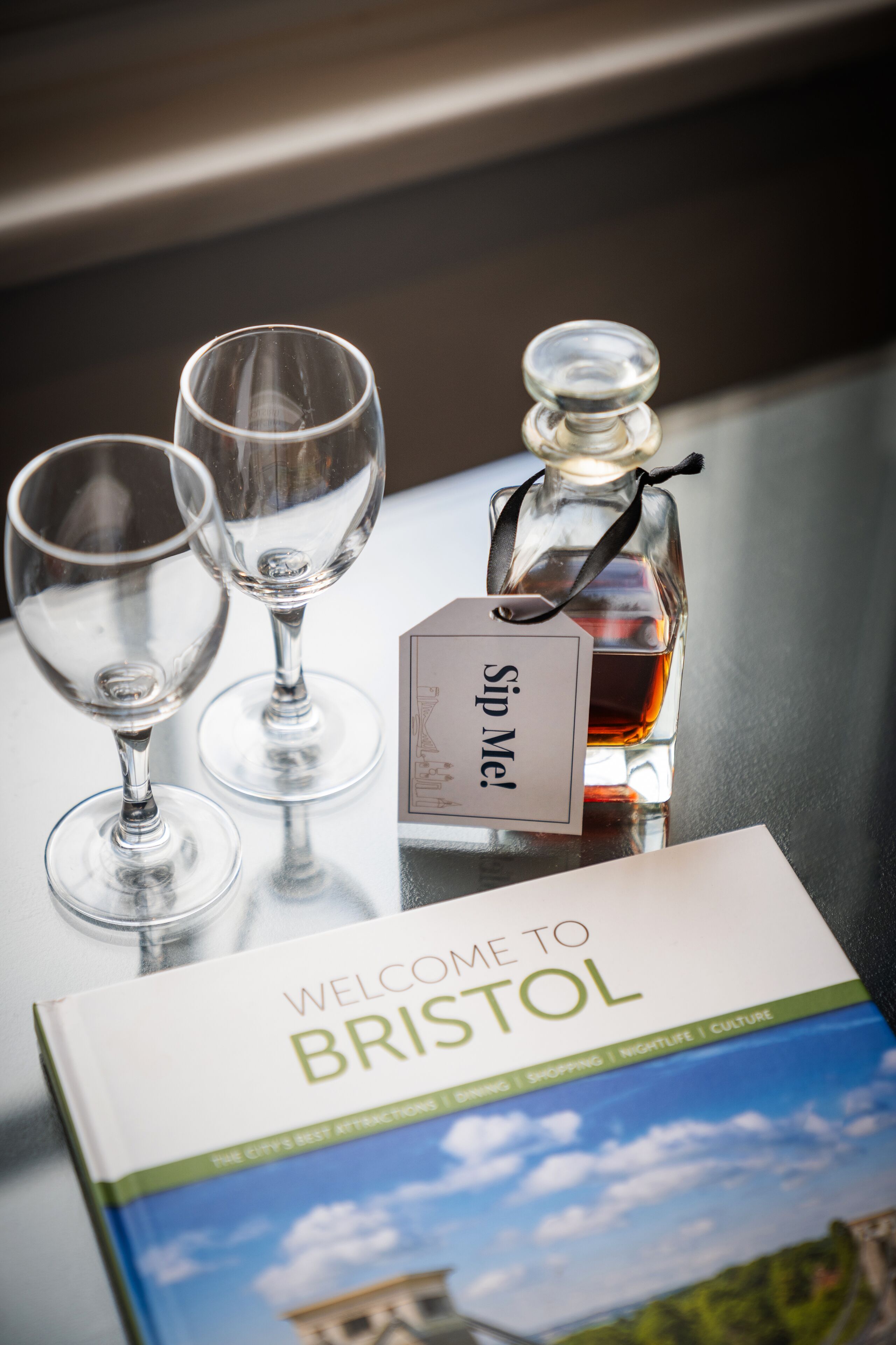 A bottle of "Sip Me!" liquor, two empty wine glasses, and a "Welcome to Bristol" brochure on a table.