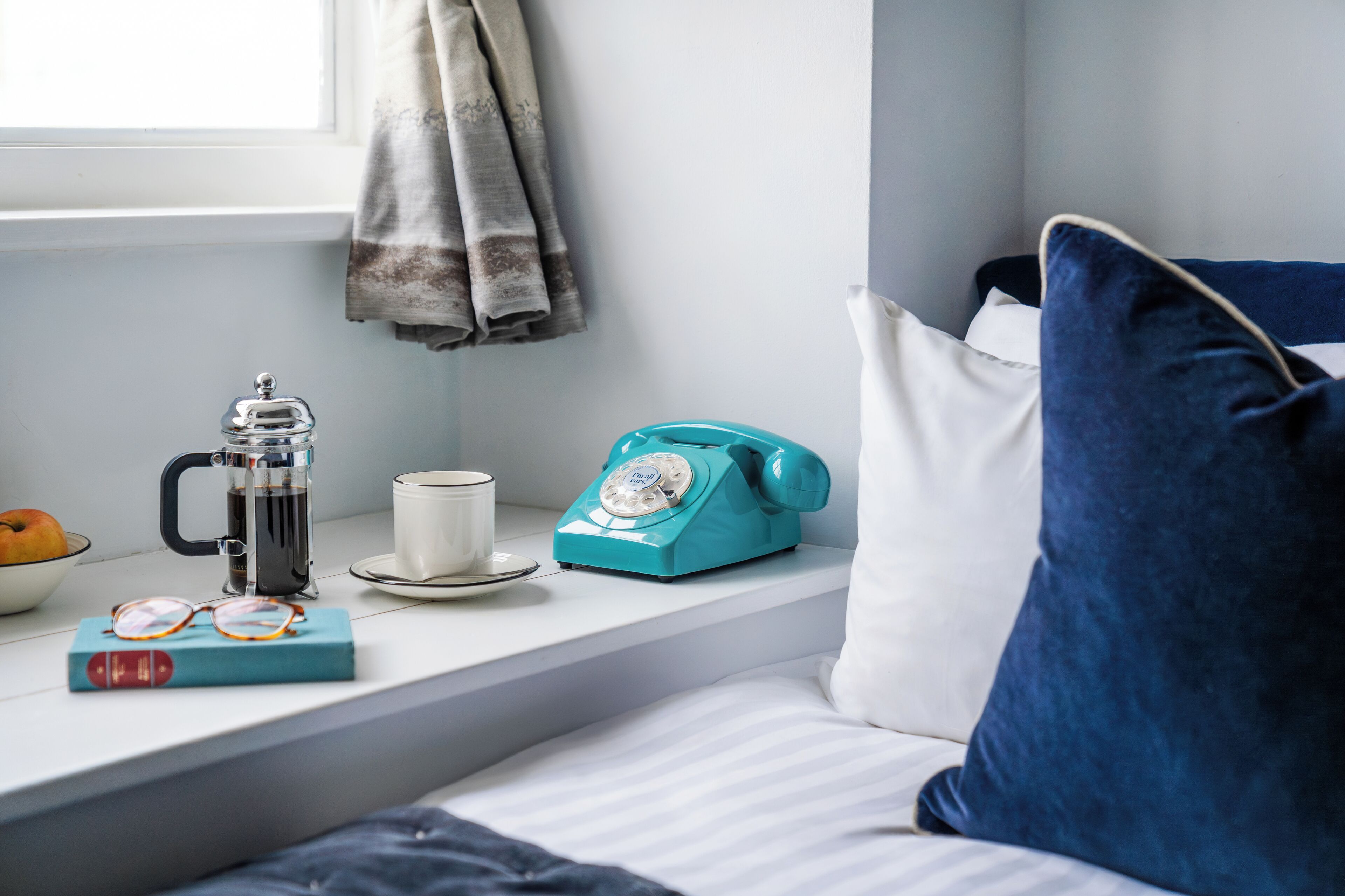 A room with a bed, a window with curtains, a coffee maker, a vintage telephone, a cup, and a book on a windowsill.