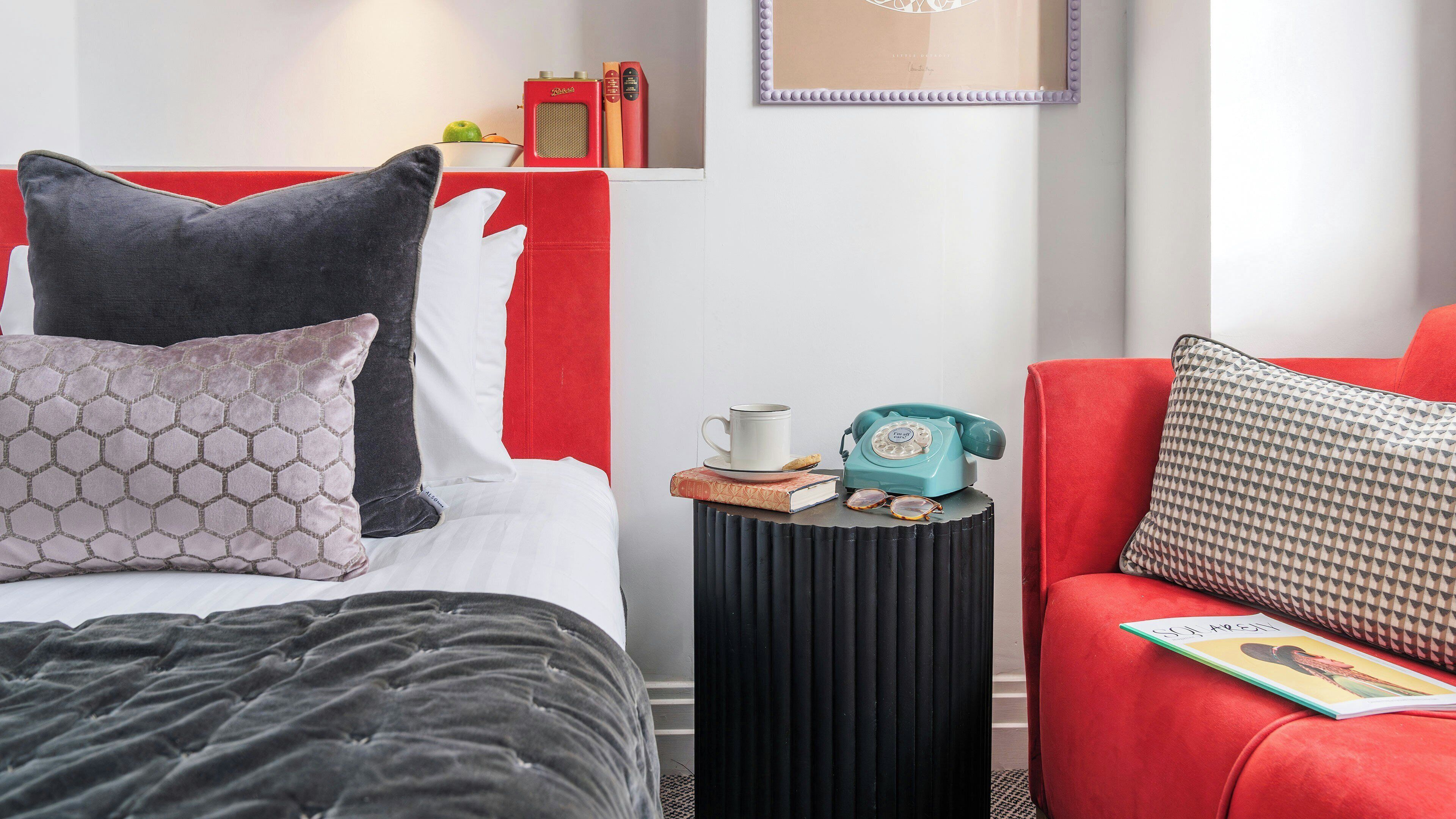 A hotel room with a bed, a red sofa, a small side table with a telephone, a framed picture on the wall, and a bookshelf with books and a fruit bowl.