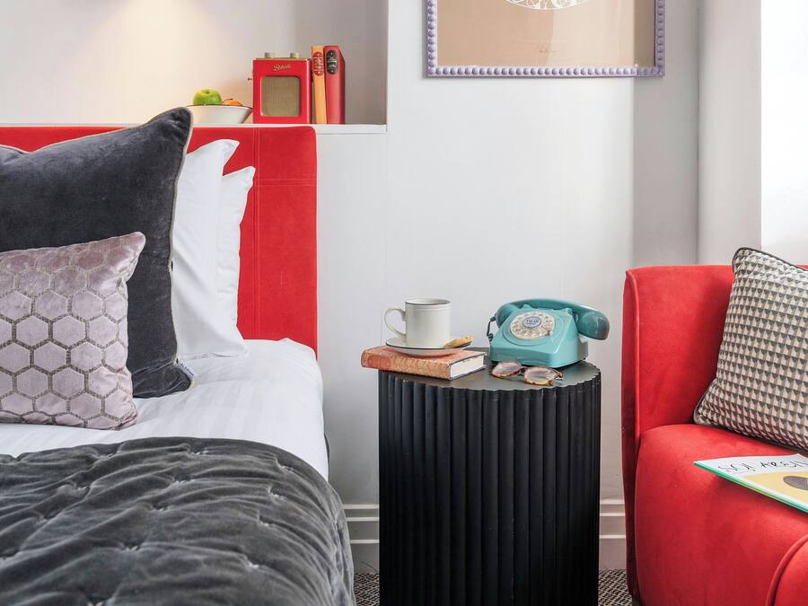 A hotel room with a bed, a red sofa, a small side table with a telephone, a framed picture on the wall, and a bookshelf with books and a fruit bowl.