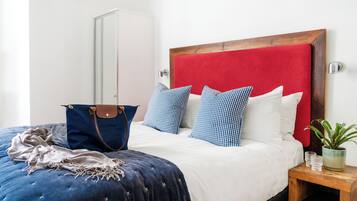 A bed with a red headboard, blue and white pillows, a blue blanket, a brown bag, and a small wooden nightstand with a plant and glasses.