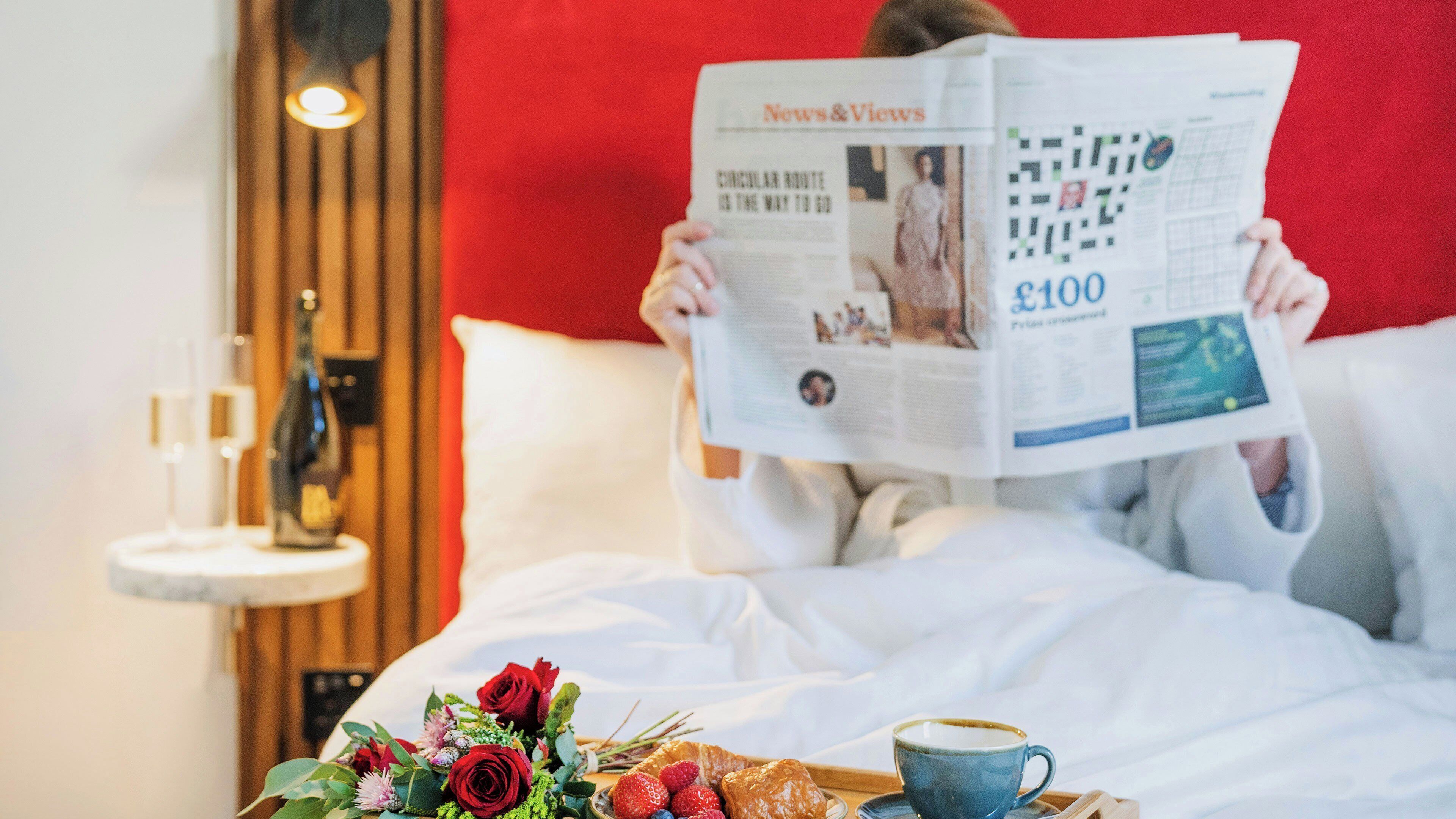 A person reading a newspaper on a bed with a tray of food and flowers.