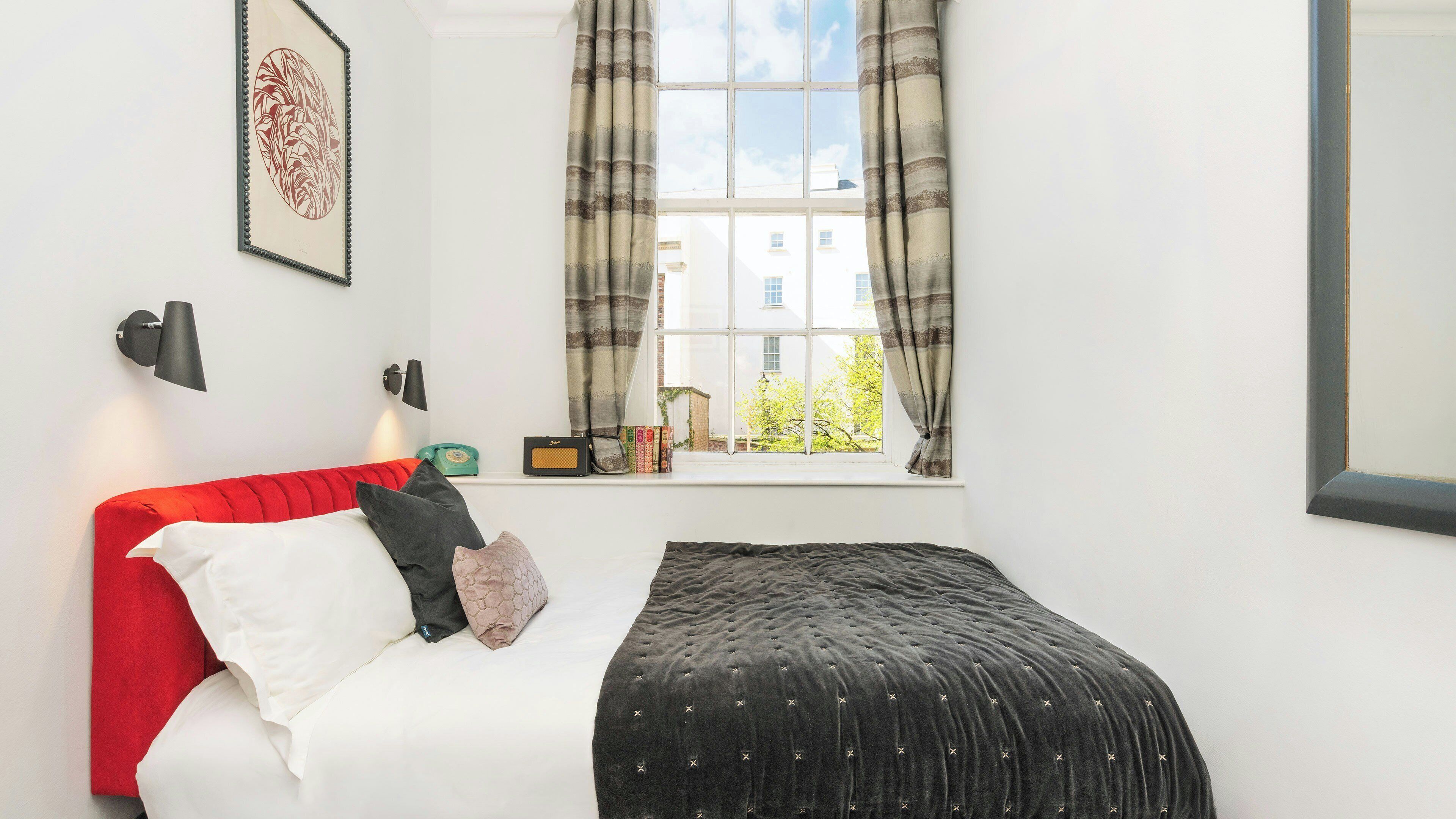 A bedroom with a red sofa, a black bed, a window with curtains, a wall-mounted lamp, and a framed picture.