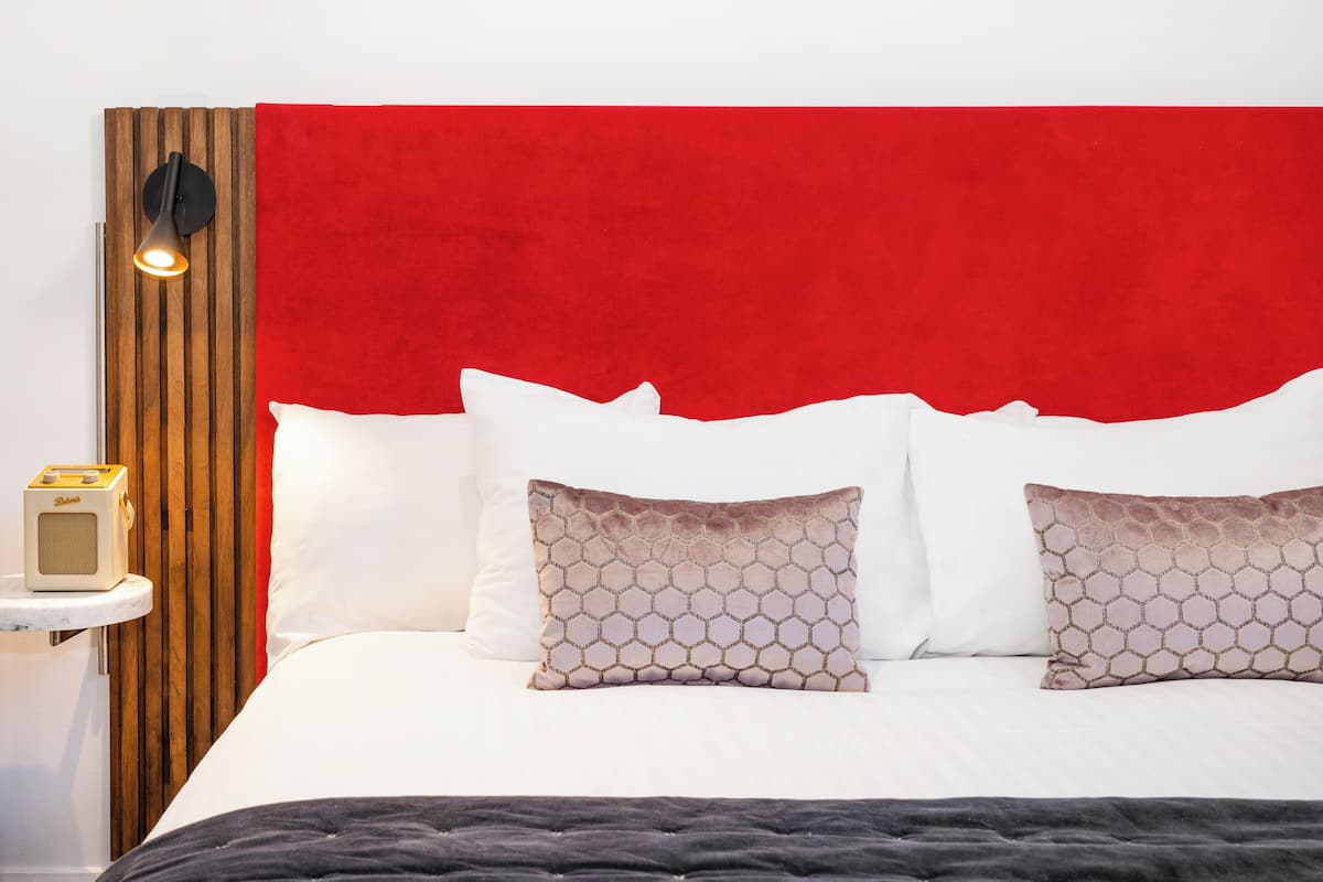 A neatly made bed with white and patterned pillows, a red headboard, and a bedside table with a lamp and a radio.