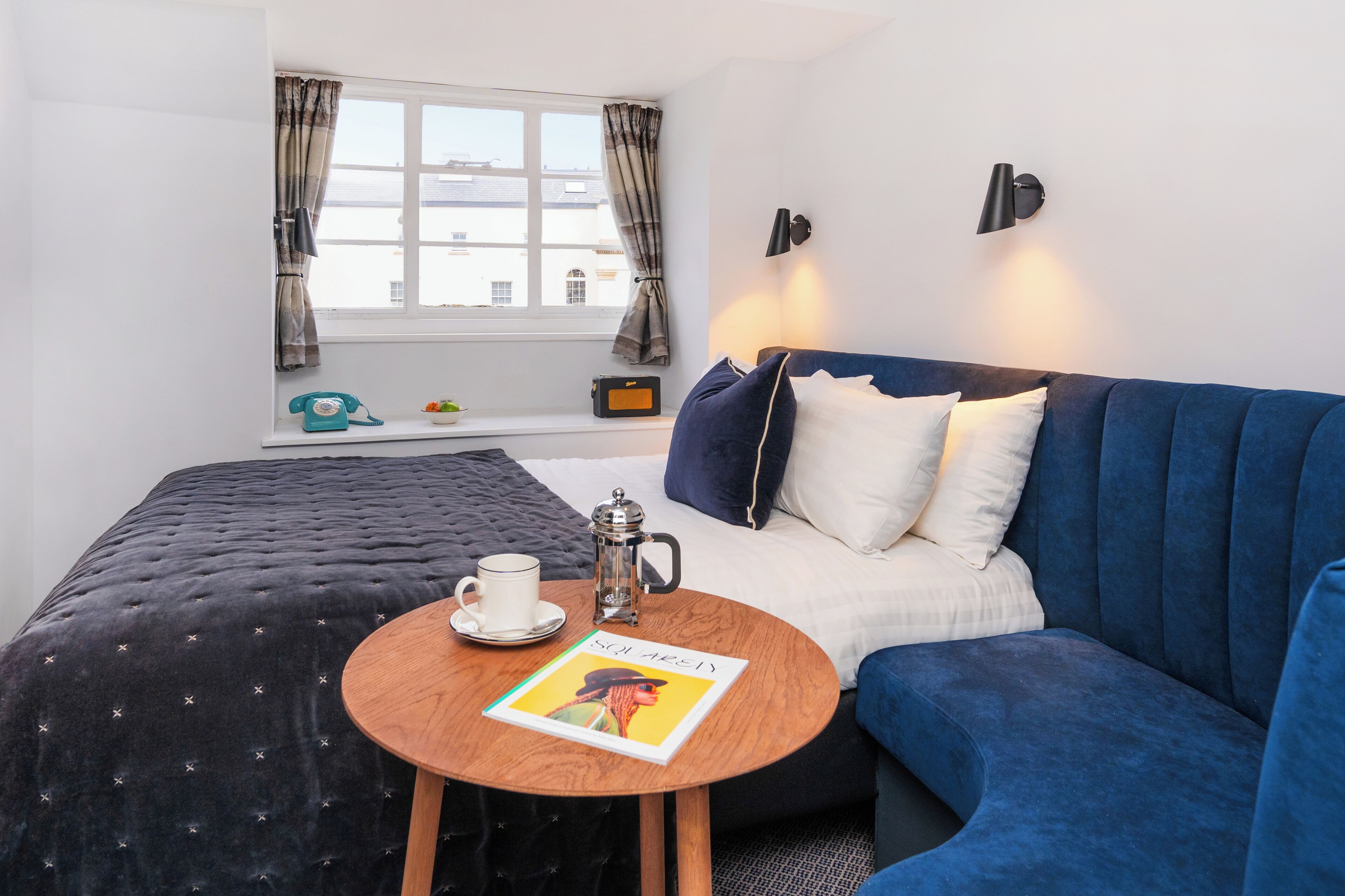 Een hotelkamer met een bed, een blauwe bank, een ronde houten tafel met een boek, een koffiezetapparaat en een telefoon.