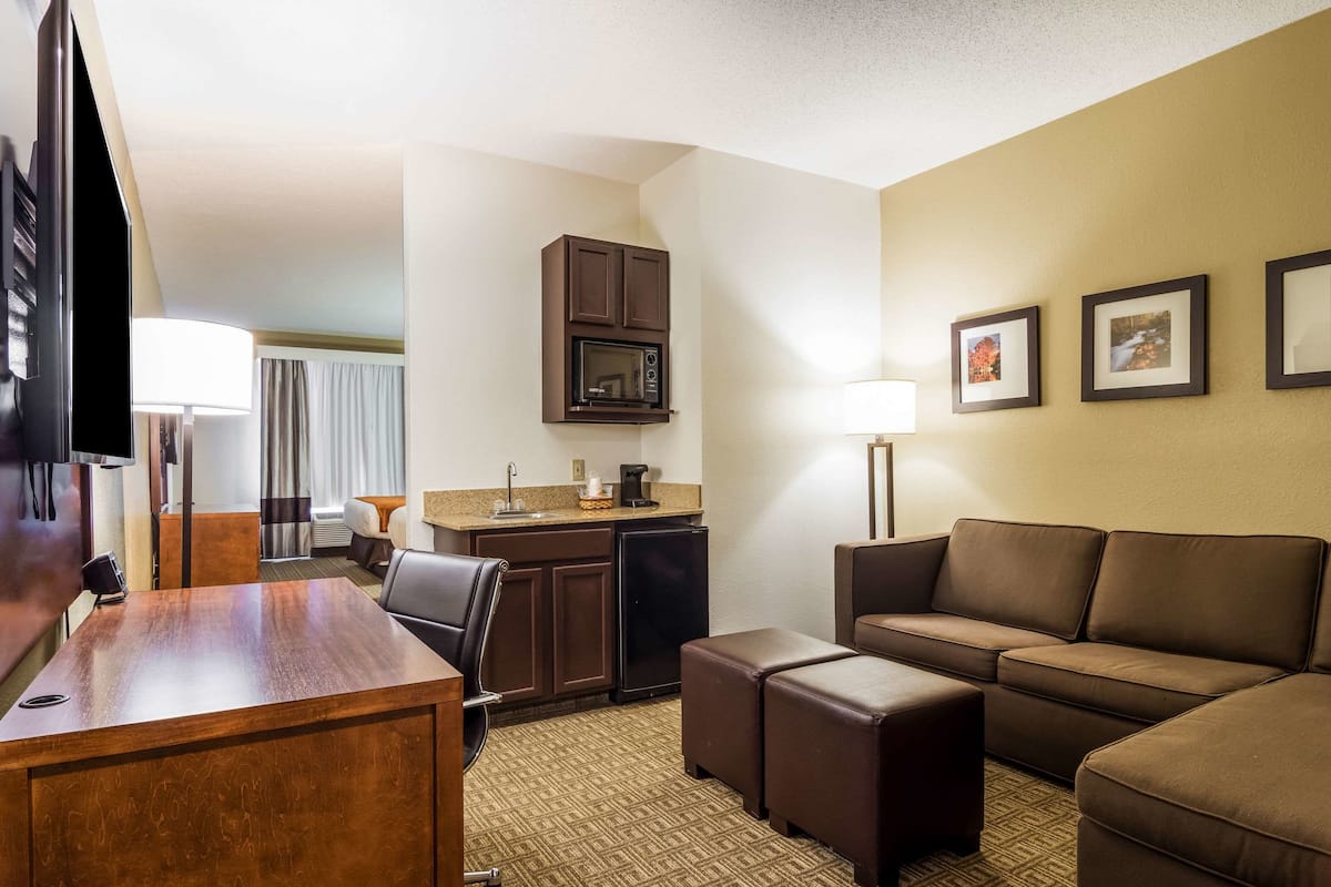 A hotel room with a wooden desk, a brown leather sofa, a small kitchenette with a microwave, and a TV.