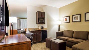 A hotel room with a wooden desk, a brown leather sofa, a small kitchenette with a microwave, and a TV.