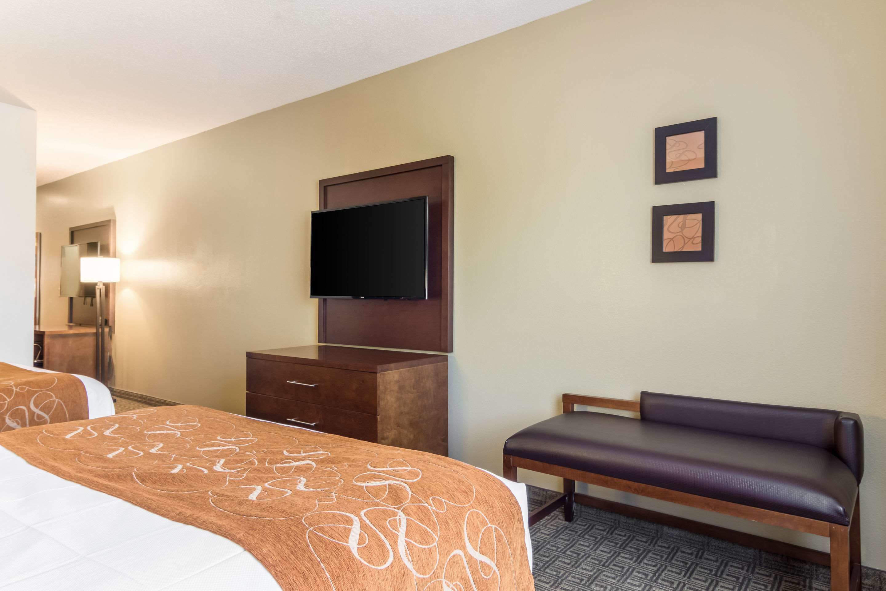 A hotel room with two beds, a flat-screen TV, a bench, and two framed pictures on the wall.