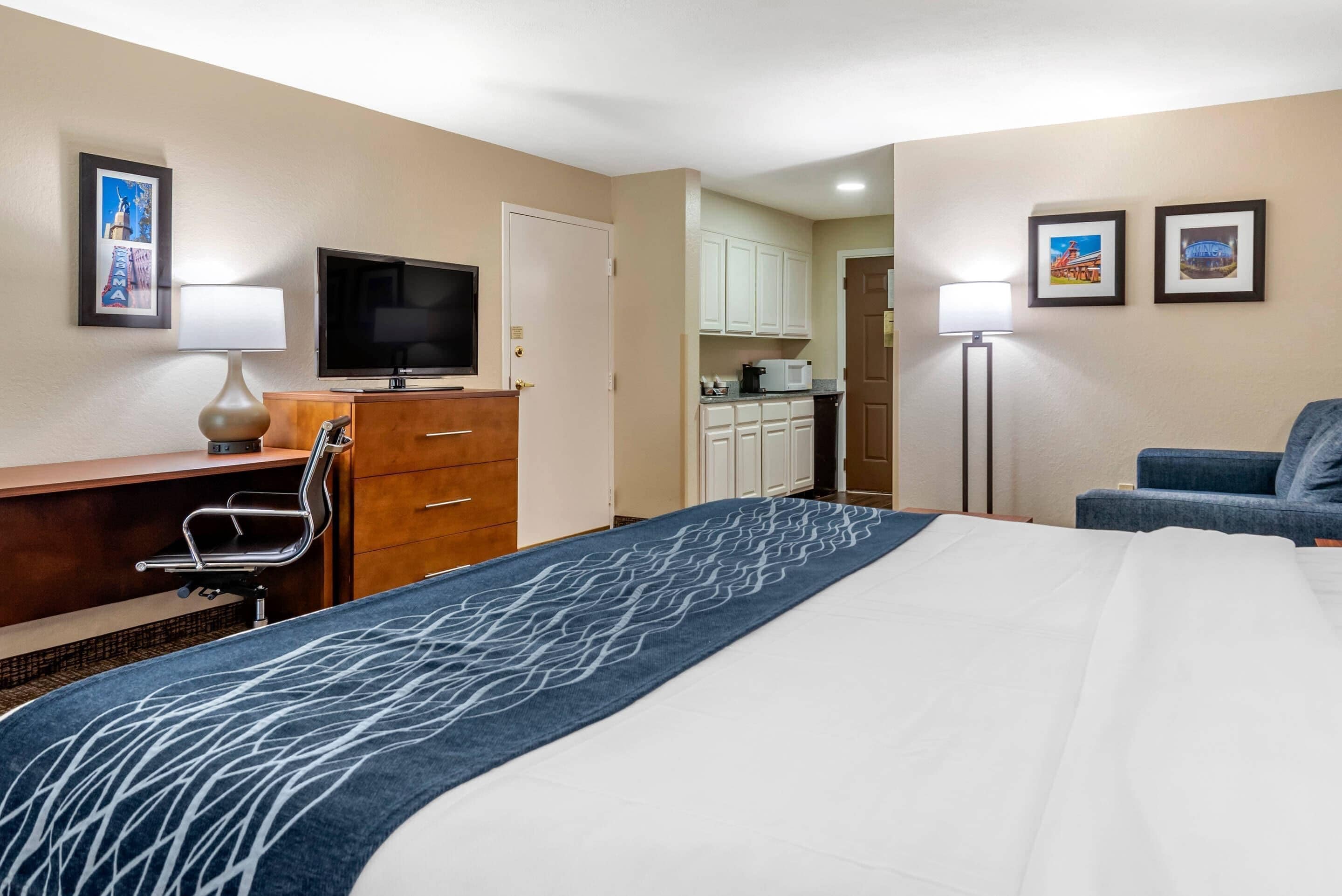 A hotel room with a bed, a desk with a chair, a television, a kitchenette, and a sofa.