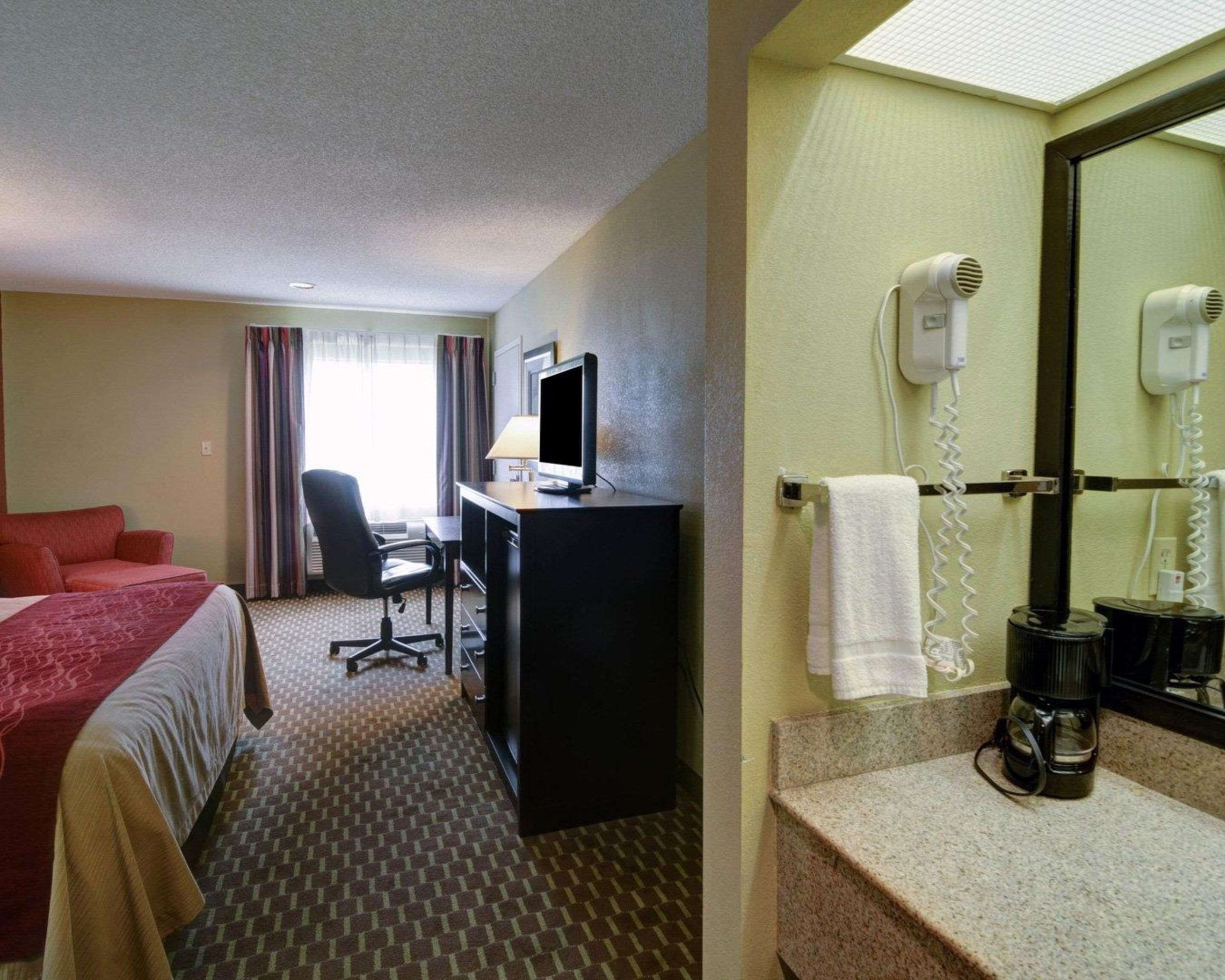 A hotel room with a bed, desk, chair, and a bathroom with a sink, mirror, and hairdryer.
