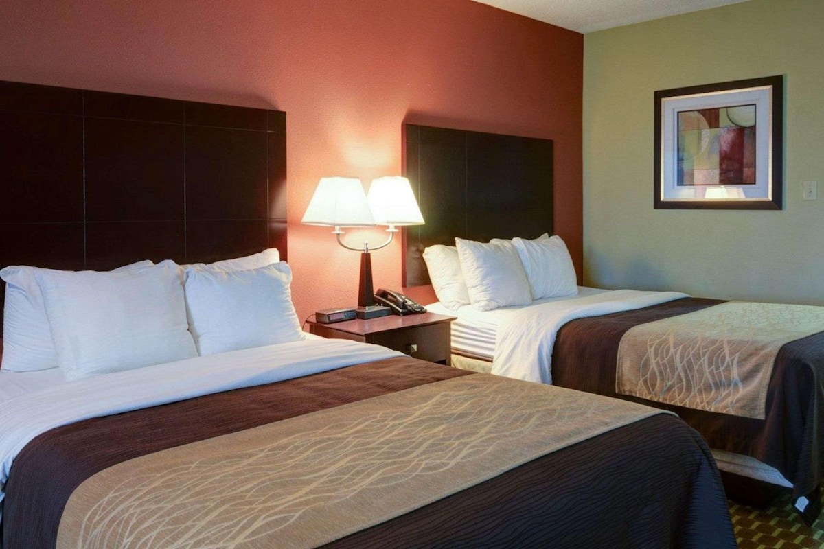 A hotel room with two beds, a nightstand with a lamp, a framed picture on the wall, and a window with blinds.