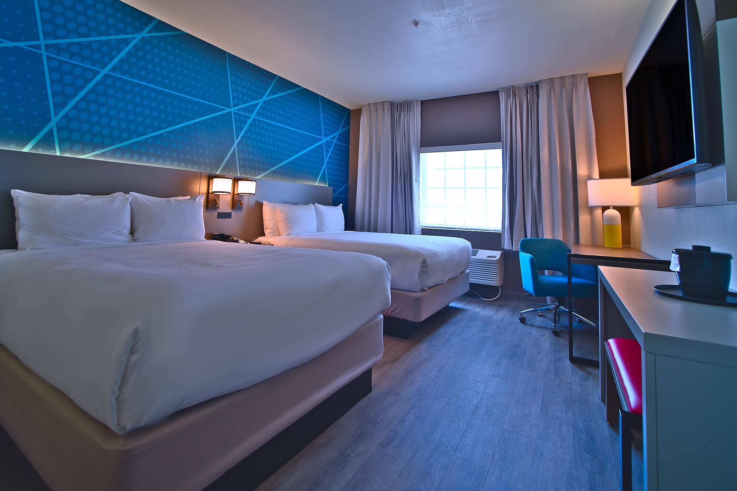 A hotel room with two beds, a desk, a chair, a TV, and a large window.