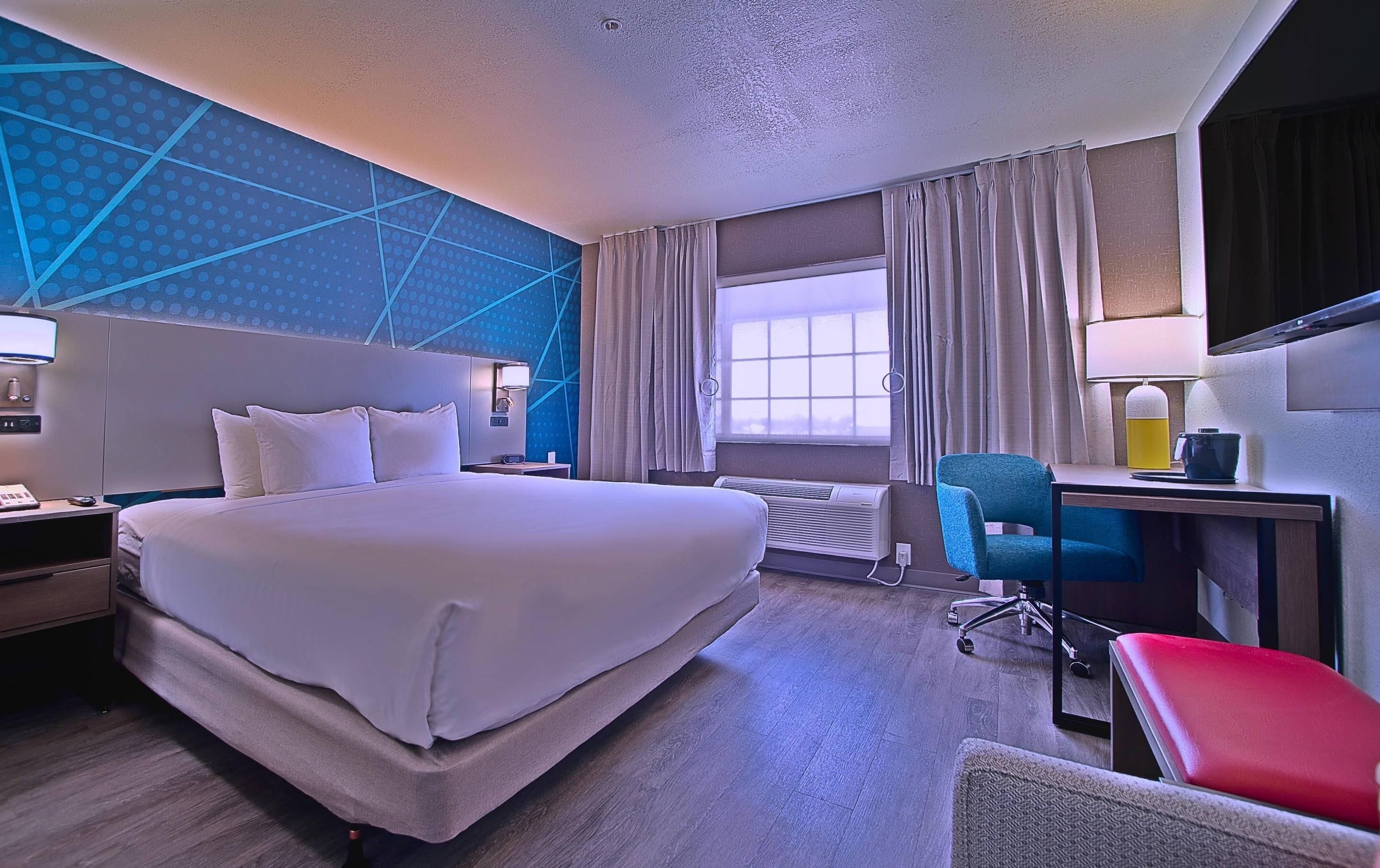 A hotel room with a large bed, a desk with a chair, a desk lamp, a telephone, and a window with curtains.