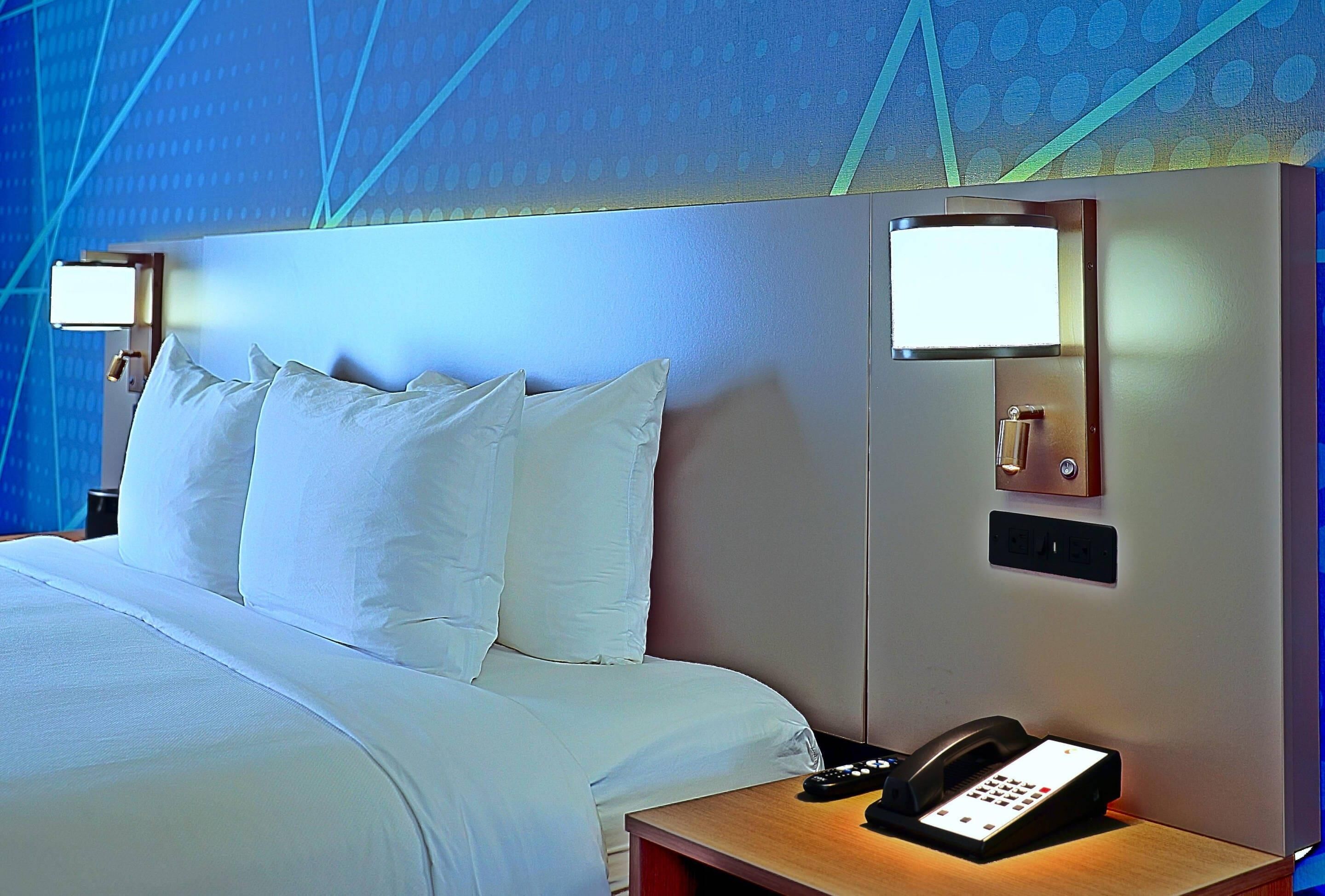 A hotel room with a bed, bedside lamps, a phone, and a wall-mounted control panel.