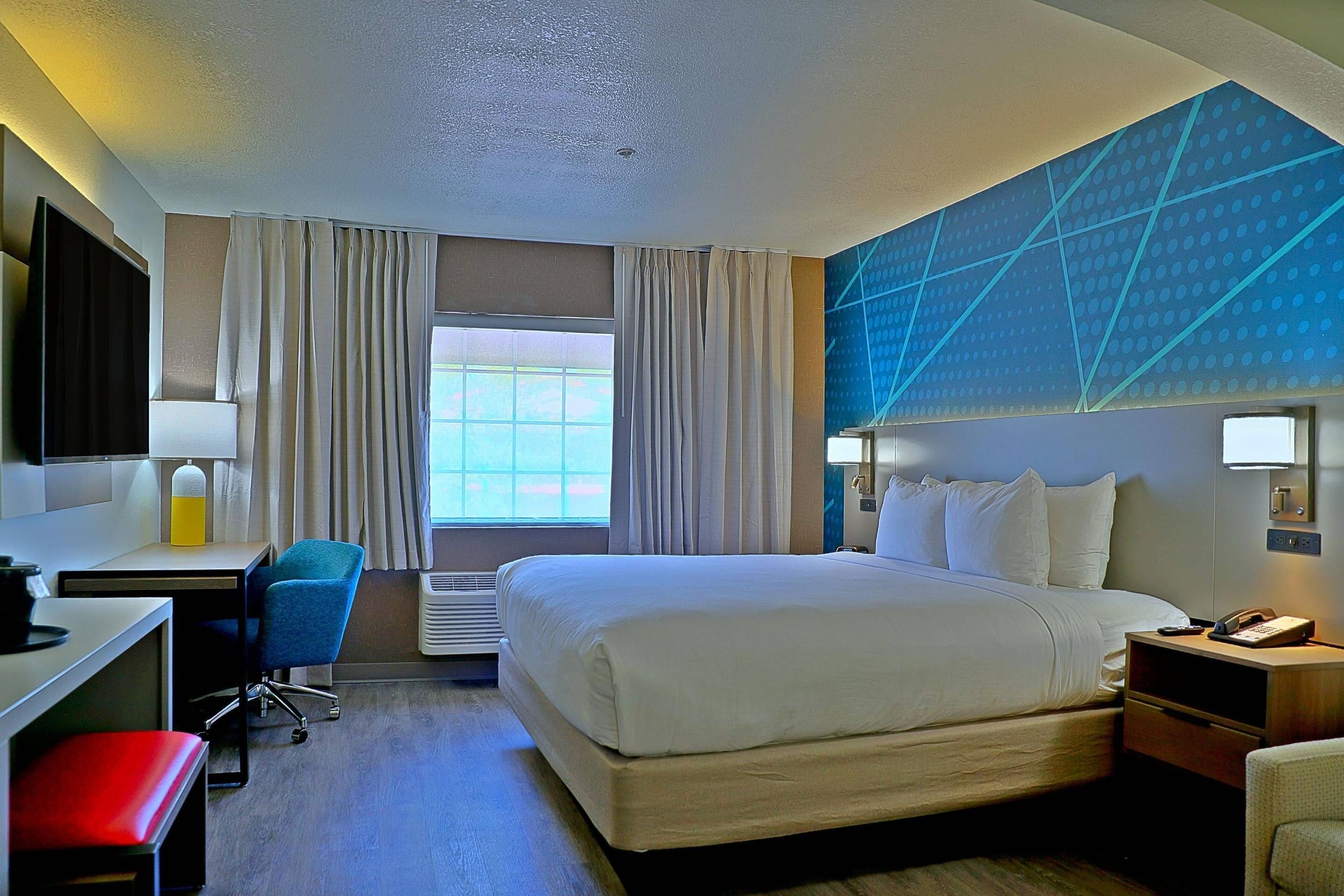 A hotel room with a bed, desk, chair, and a large window with curtains.