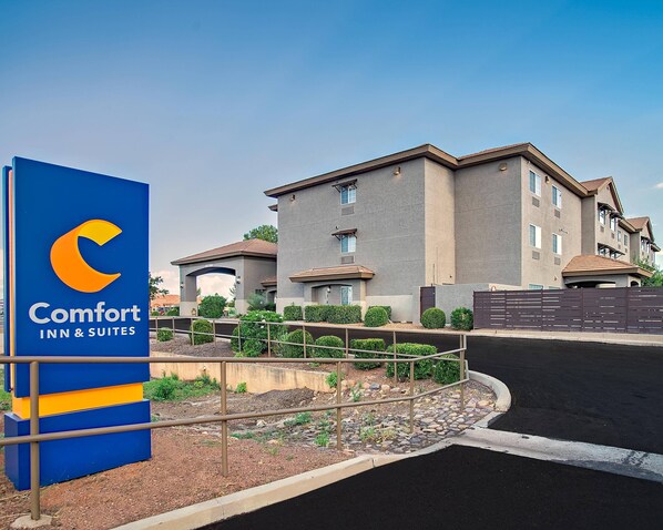 Edificio de Comfort Inn & Suites con un letrero azul, entrada con portón y un área ajardinada con arbustos y un pequeño jardín.