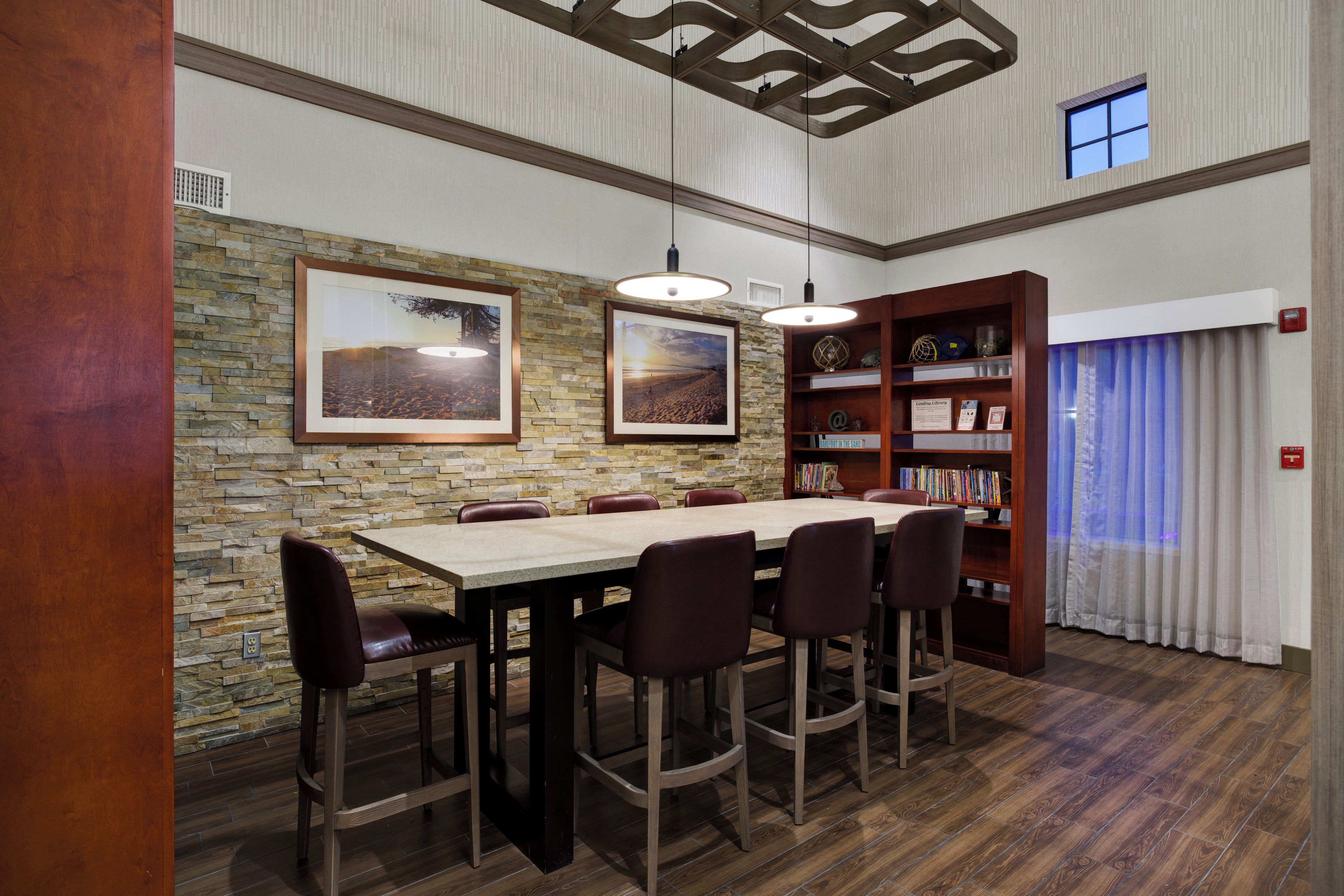 Un comedor con pared de piedra, mesa de madera y sillas. Se ve una estantería con libros y objetos decorativos.
