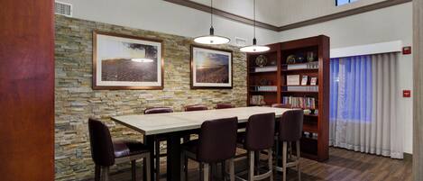 Un comedor con pared de piedra, mesa de madera y sillas. Se ve una estantería con libros y objetos decorativos.