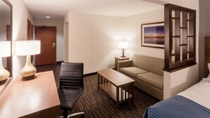 A hotel room with a bed, a sofa, a desk with a lamp, a chair, and a picture on the wall.