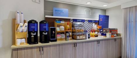A hotel breakfast area with coffee machines, a bakery display, and a sign indicating breakfast is ready.