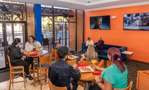 Un grupo de personas sentadas en mesas en una sala con una pantalla grande mostrando un noticiero.