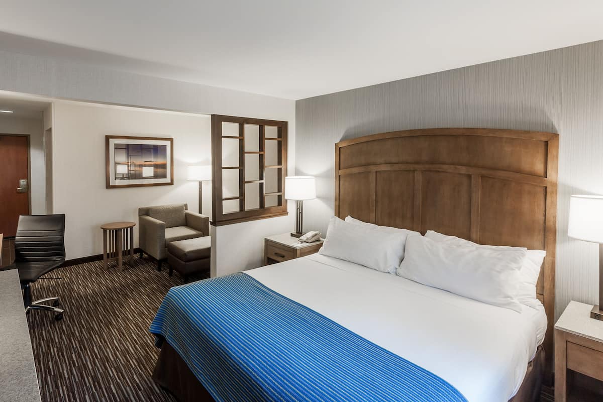 A hotel room with a large bed, a desk, a chair, a small table, and a nightstand with a lamp.