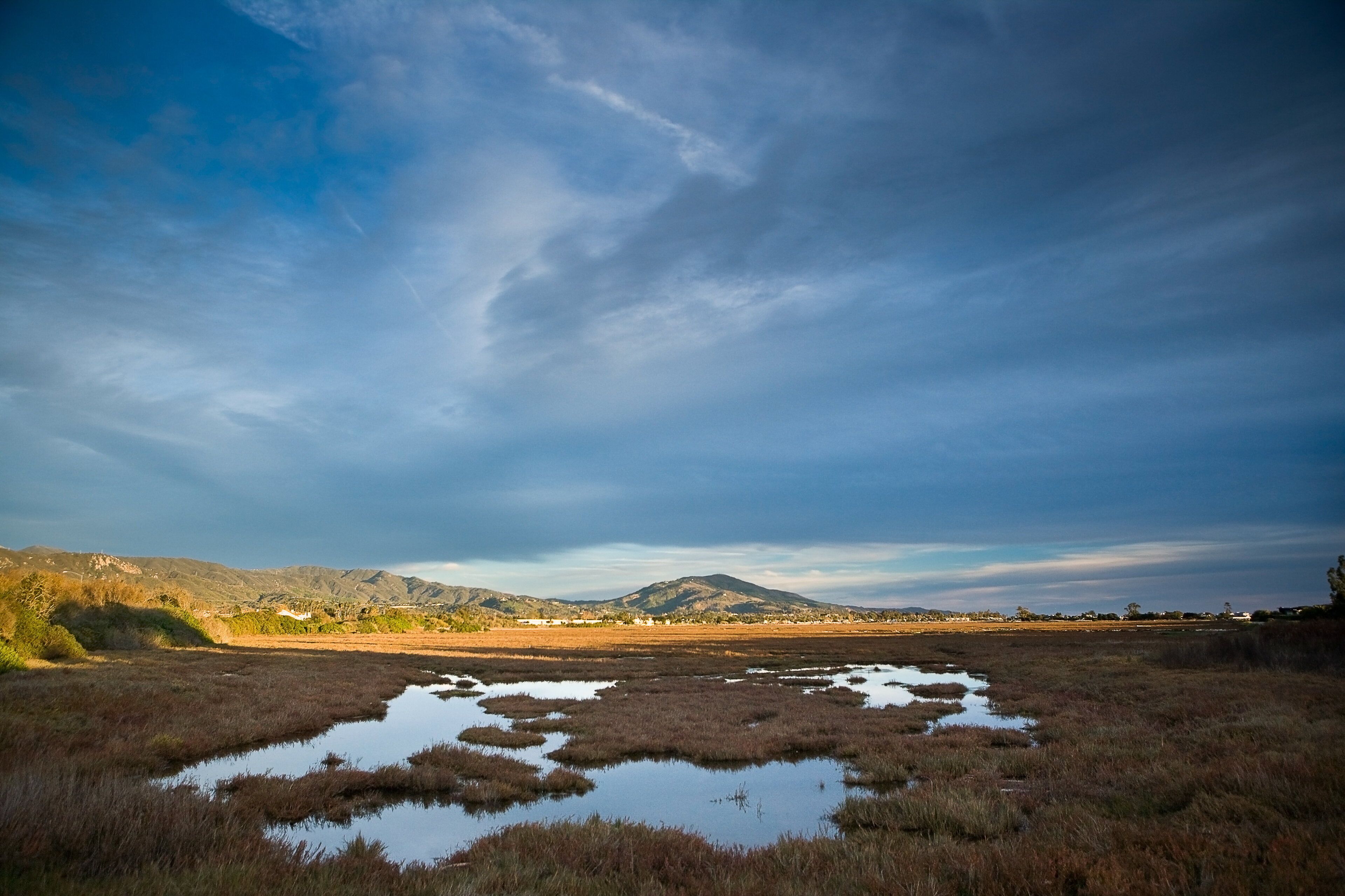 A natural landscape with a flat terrain, water bodies, and distant mountains under a cloudy sky.