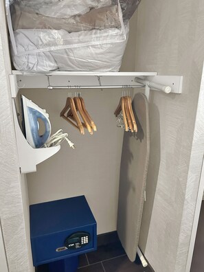 A storage closet with hanging wooden hangers, a white ironing board, and a blue storage box with a control panel. - Holiday Inn Express & Suites Carpinteria by IHG (Carpinteria)