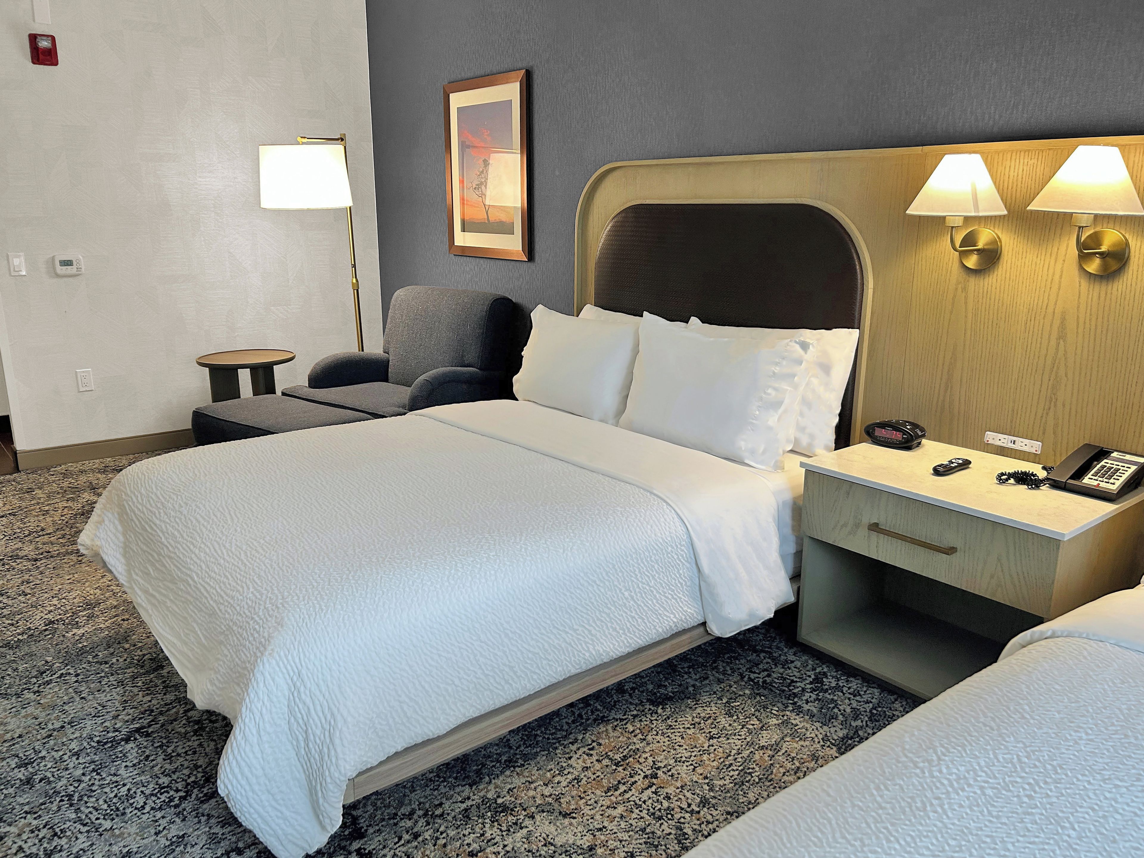 A hotel room with a bed, bedside table, lamp, and a painting on the wall.