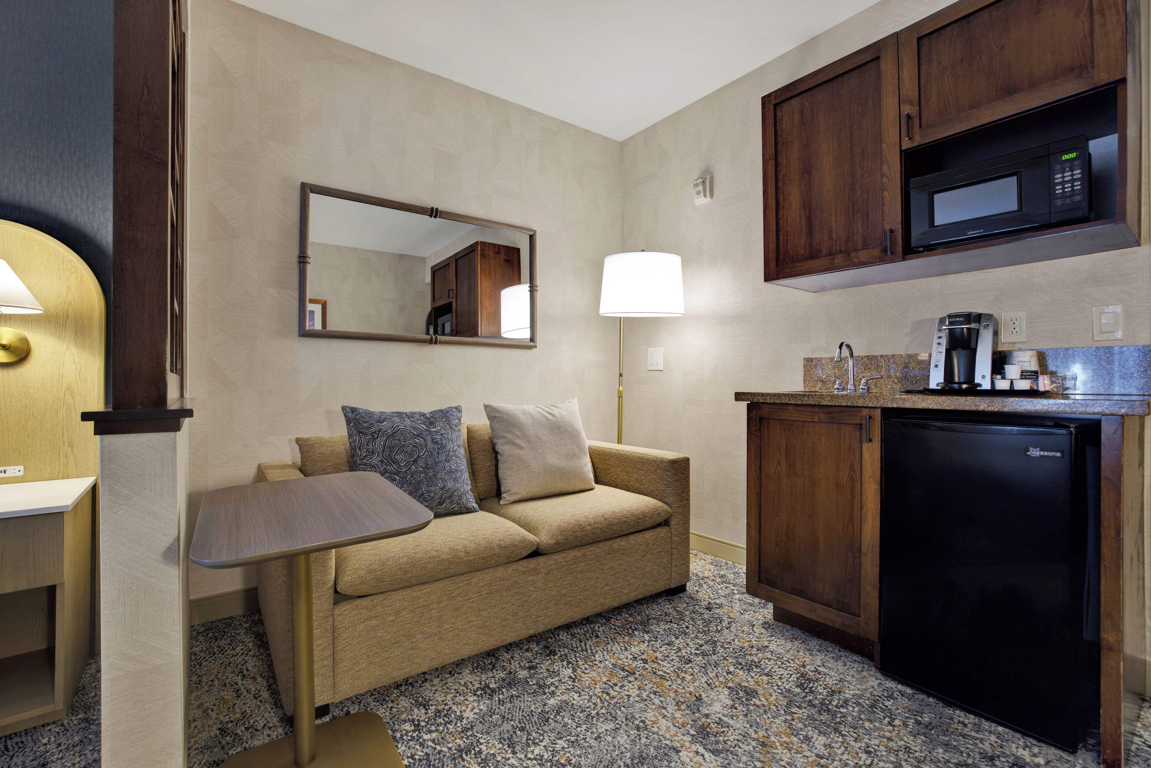 Ein gemütliches Hotelzimmer mit Küchenzeile, einer Couch mit Kissen, einem kleinen Tisch, einer Lampe und einer Mikrowelle.