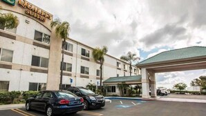 A hotel with a sign reading "Quality Suites," featuring a parking lot with several cars, a covered parking structure, and palm trees. - Quality Suites San Diego Otay Mesa (San Diego)