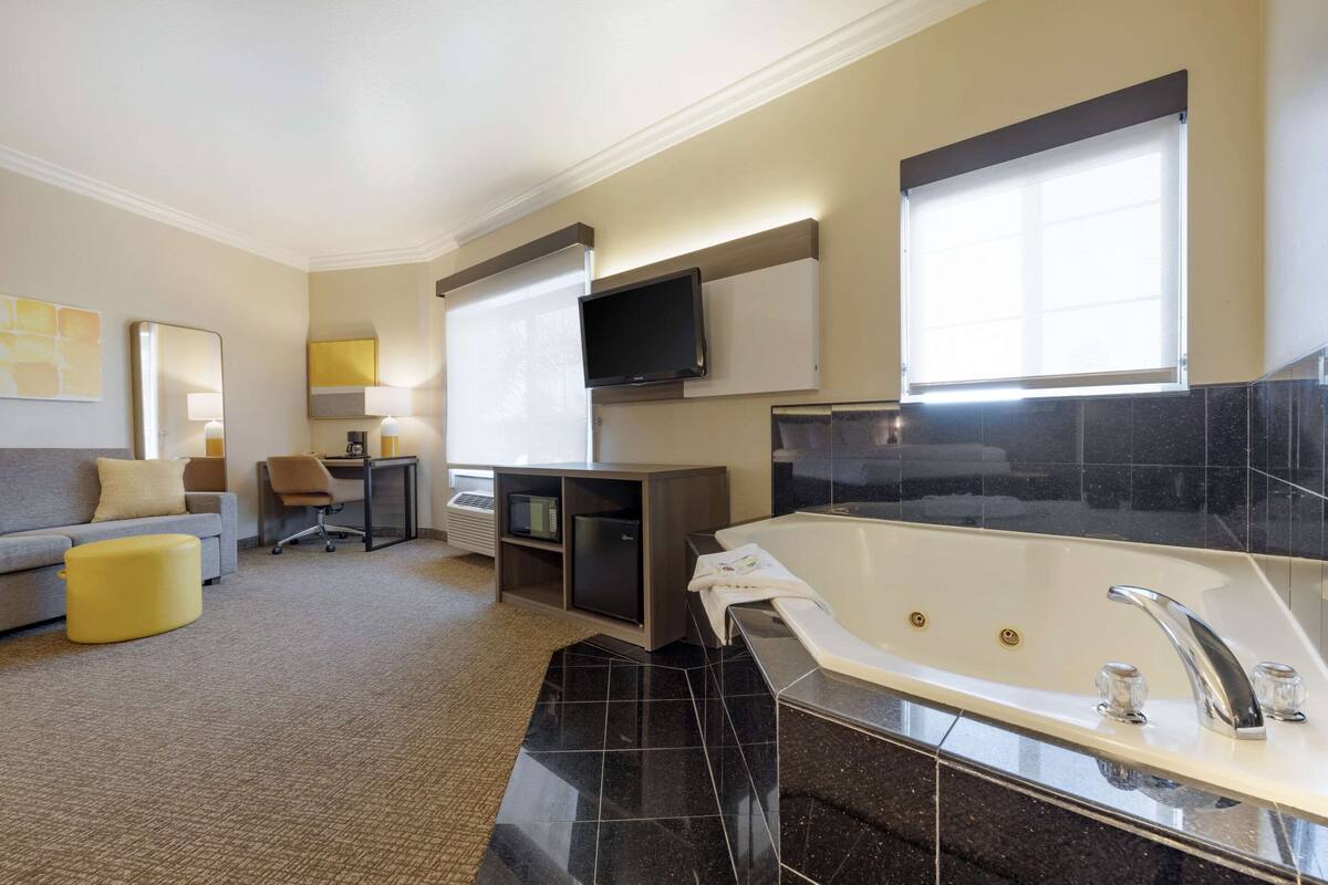 A modern hotel bathroom with a large bathtub, a vanity with a mirror, and a flat-screen TV mounted on the wall.