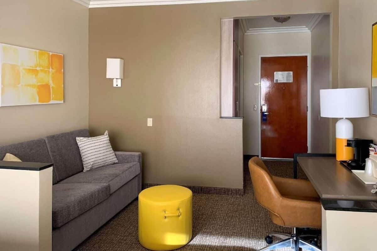 A hotel room with a sofa, a yellow ottoman, a desk with a lamp, and a door leading to another room.