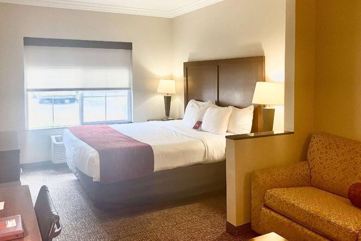 A hotel room with a bed, two bedside lamps, a sofa, a desk with a chair, and a window with blinds.