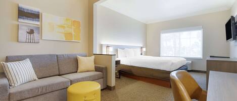 A hotel room with a bed, a sofa, a small yellow ottoman, and a desk.
