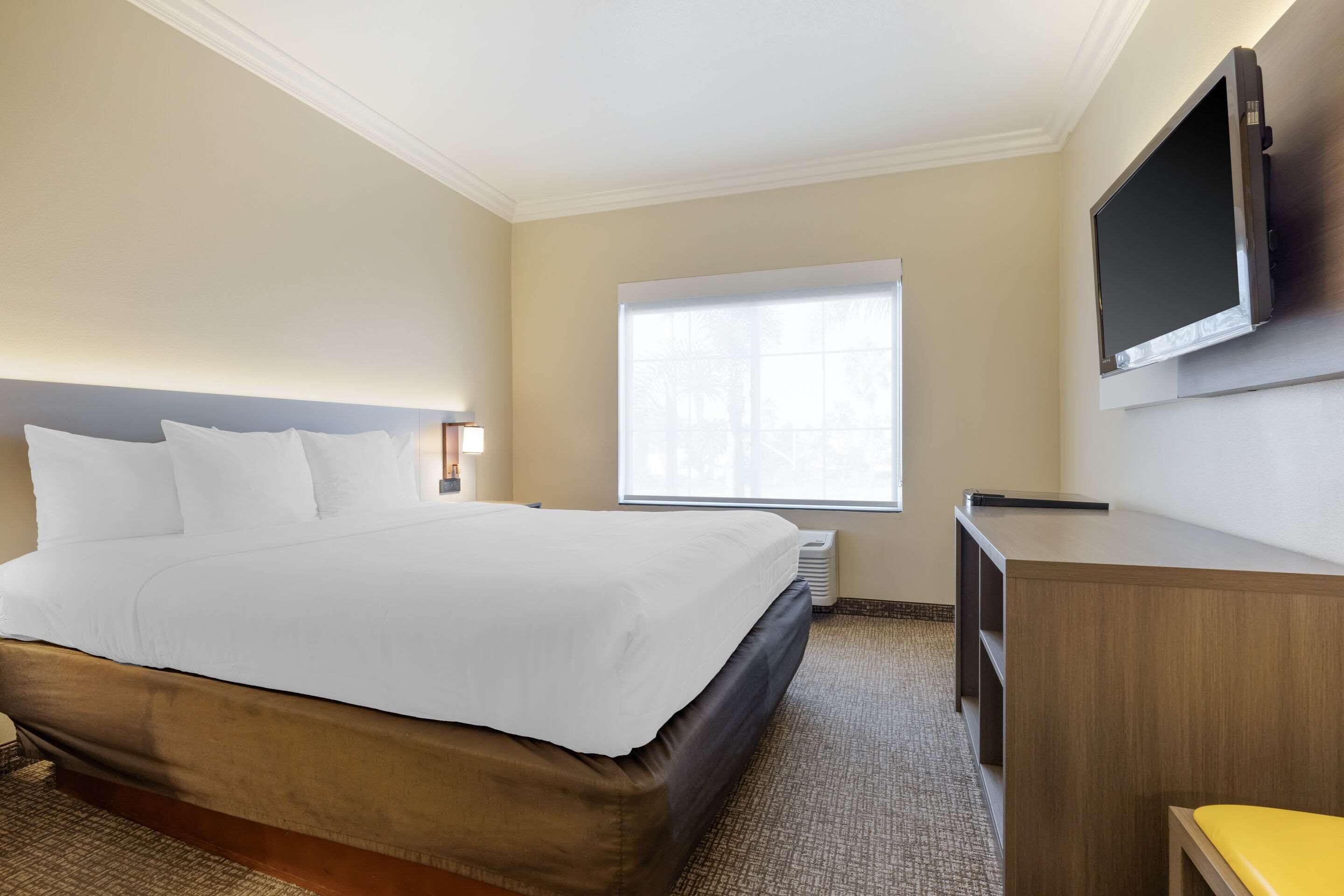 A hotel room with a large bed, a flat-screen TV mounted on the wall, a window with blinds, and a small desk with a chair.